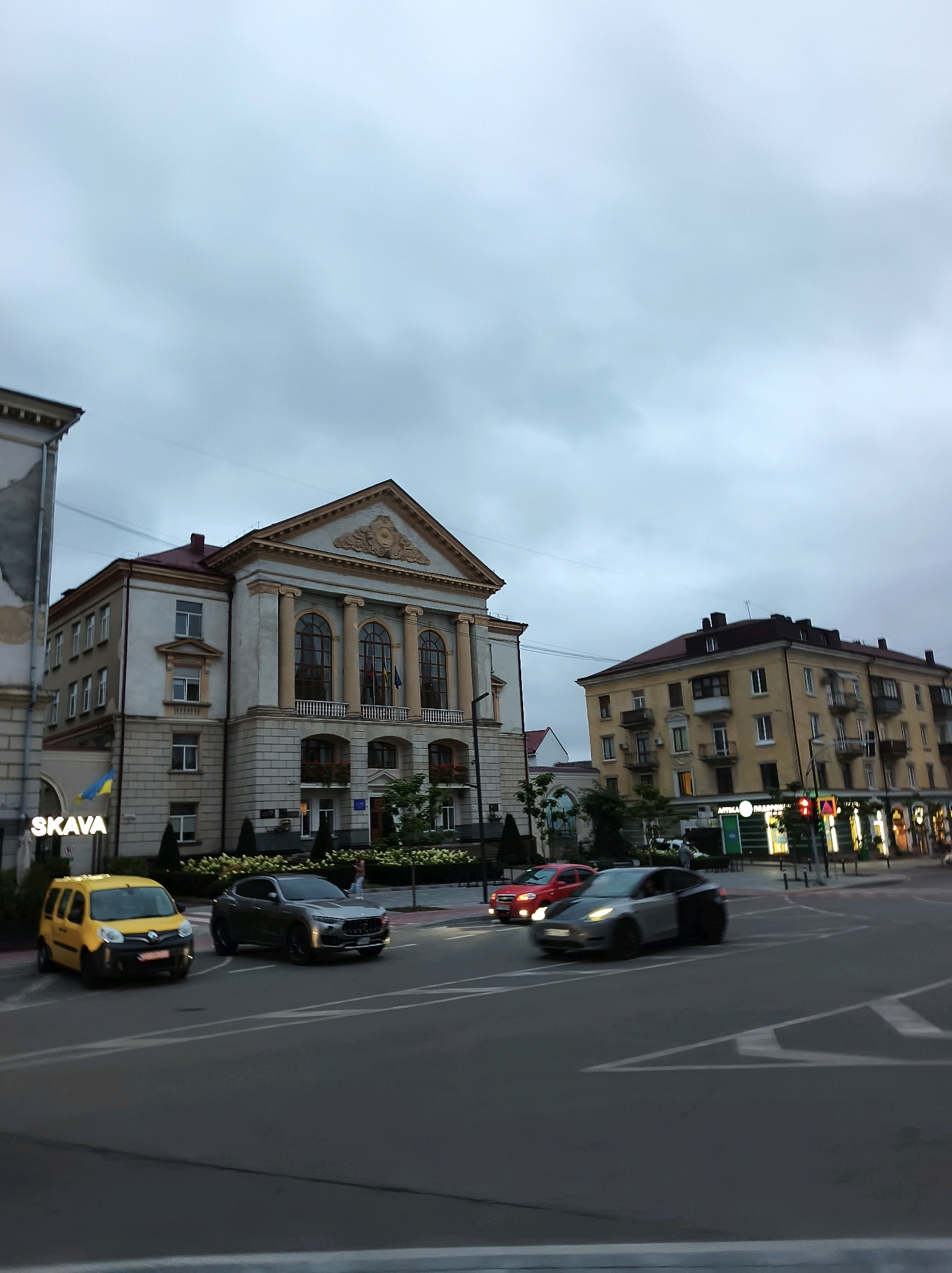 The image captures a view of the Ternopil Regional Drama Theatre, a significant architectural landmark in Ternopil, Ukraine. The theatre, with its grand facade featuring columns and arched windows, is prominently displayed. In front of the theatre, a yellow car and a silver SUV are visible on the road. The sky is overcast, suggesting a cloudy day. To the right of the theatre, a yellow apartment building with several windows and balconies is also present. The street in the foreground shows road markings, and several other vehicles are seen driving or parked. The overall scene depicts a street view in a Ukrainian city, with the theatre as the central point of interest.