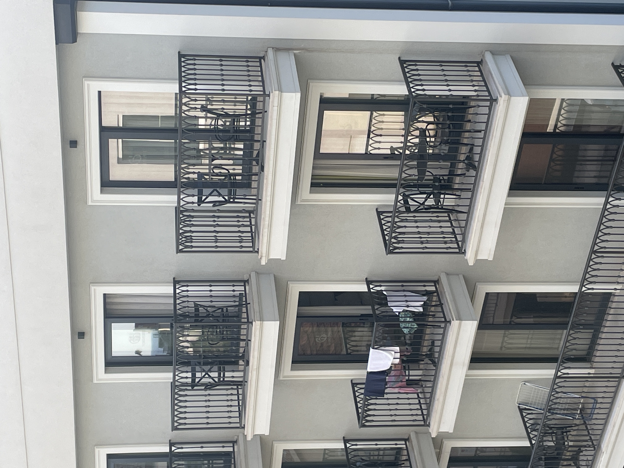 The image shows the facade of an apartment building. The building is several stories tall, and each apartment has a balcony with black iron railings. Some of the balconies are empty, while others have furniture or clothes hanging on them. The building is white or light gray. The scene is in Budva, Montenegro.