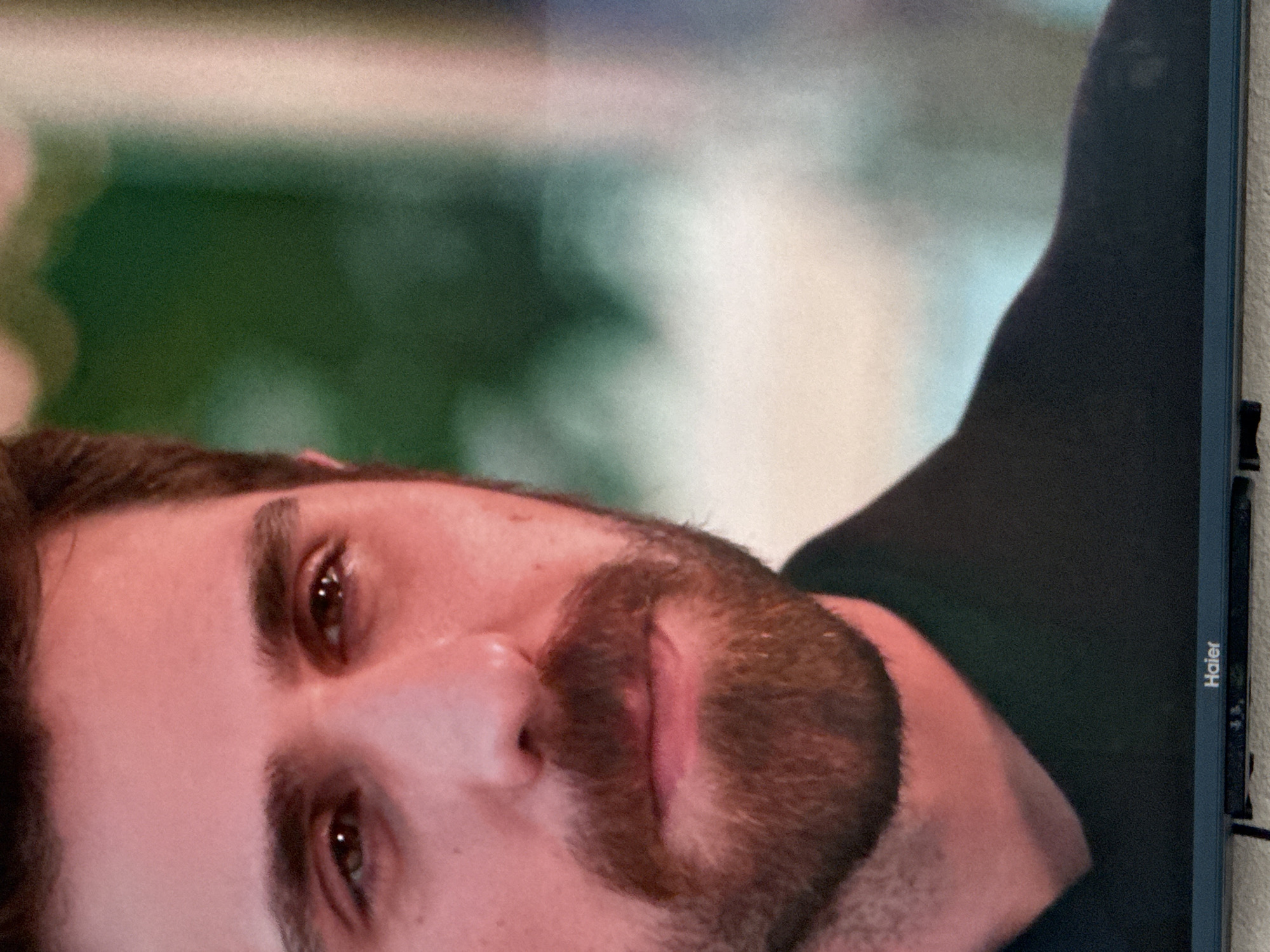 The image shows a close-up of a man's face. He has dark hair and a beard. His eyes are brown, and he has a slight smile on his lips. The background is out of focus, but it appears to be an outdoor setting with greenery. There is a television screen with the brand "Haier" visible at the bottom of the image. The man is wearing a dark shirt. The lighting suggests an indoor setting or a time of day with warm light. The location of Uyutnoye, Ukraine, is not discernible from the visual information provided in the image.
