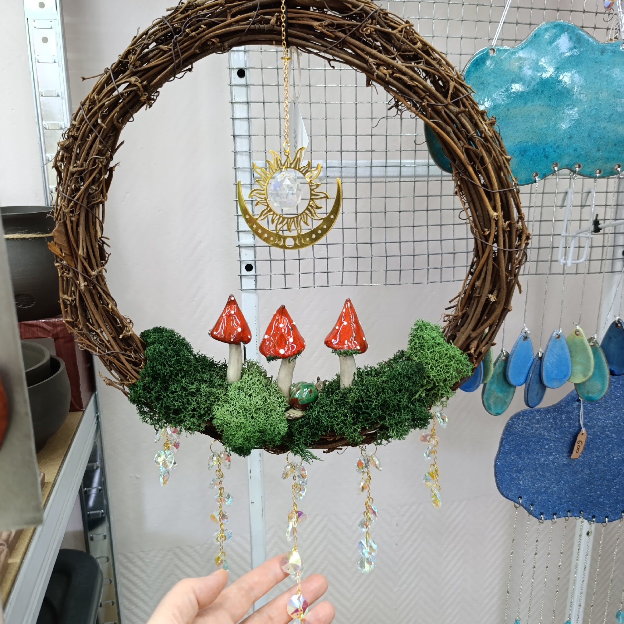 A handmade, decorative wreath featuring natural and artificial elements hangs against a wire mesh display. The wreath is constructed of interwoven brown twigs, forming a circle. Attached to the bottom is a bed of vibrant green moss, from which three ceramic mushrooms sprout, each with a red, glossy cap and white spots. A small, glazed ceramic snail sits amidst the moss. Below the moss, several strands of crystalline prisms hang, catching the light. A gold-colored sun and moon ornament hangs from the top center of the wreath. In the background, other artisanal items are displayed, including a blue ceramic cloud and teardrop-shaped ornaments. A hand is partially visible in the foreground. The scene appears to be a craft fair or artisan market display, likely in Chaikovsky, Russia.