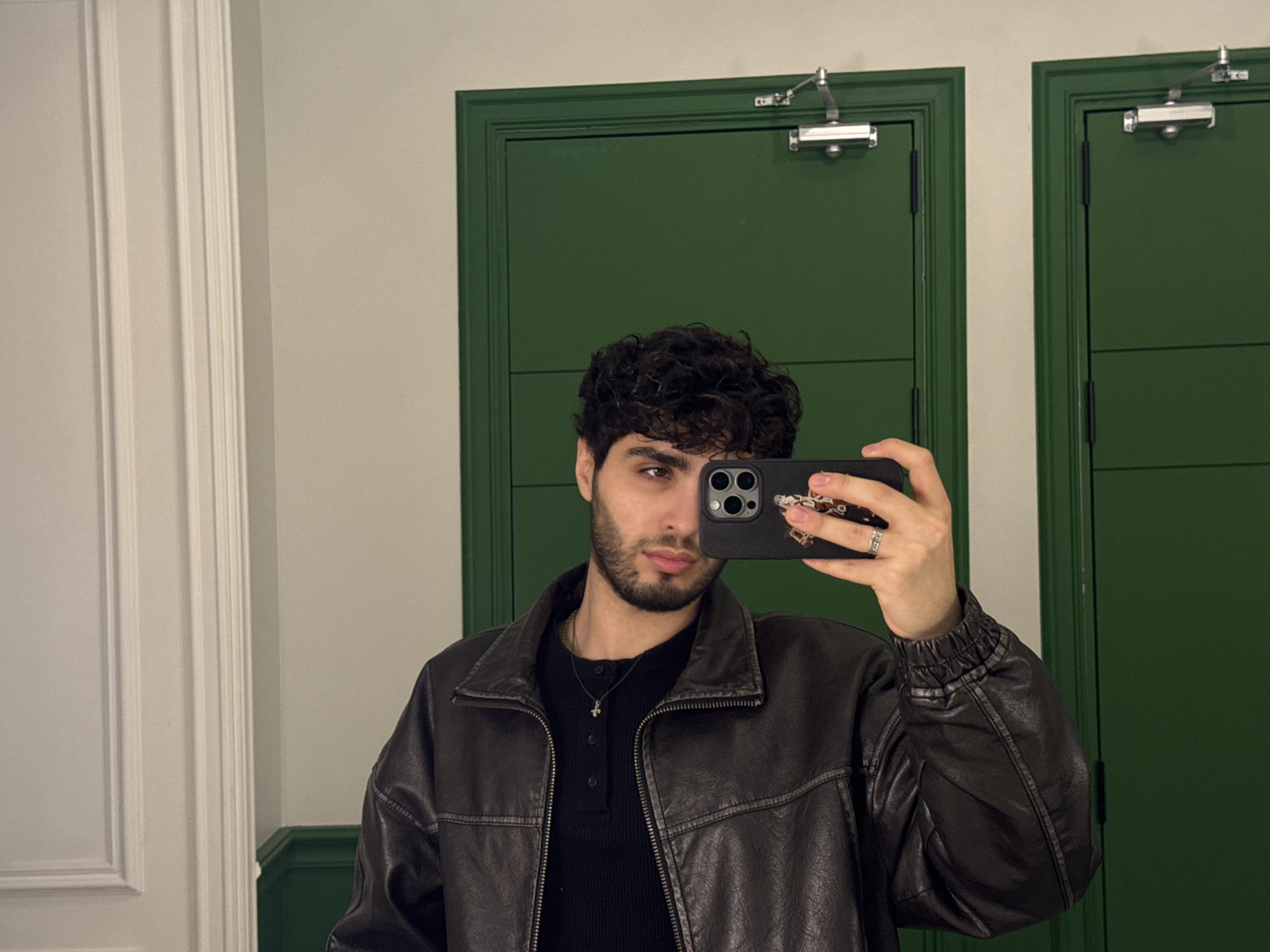 A young man takes a selfie in a room with green doors. The man has dark curly hair, a beard, and is wearing a black shirt under a brown leather jacket. He holds a smartphone with a black case up to his face, obscuring one eye. Behind him are two green doors with silver hardware. To the left is a white paneled wall. The image provides no information about the location being in Yerevan, Armenia.