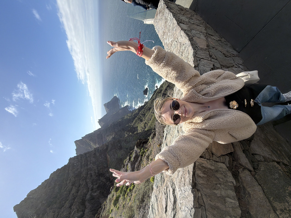 An outdoor scene depicts a female subject posing at a coastal viewpoint in the city of Tirma, Spain. The subject, a Caucasian female with light-colored hair pulled back, is wearing oval-shaped sunglasses, a beige teddy fleece jacket, a black strapless top with three gold-colored decorative clasps, light blue jeans, and white sneakers. She is smiling towards the viewer and has both arms raised above her head with open palms. A light pink string bracelet is visible on her left wrist, while her right wrist displays multiple red and white string bracelets. A small, white, quilted handbag with a chain strap hangs on her left side, partially obscured by a stone wall.
The subject is positioned behind a multi-toned grey stone wall, constructed from irregular blocks, which forms a barrier for the viewpoint. To the far right, a transparent glass barrier and the partial figure of another individual in dark clothing are visible behind the stone wall.
The background consists of dramatic, dark grey-brown volcanic cliffs with sparse green vegetation, descending steeply into a deep blue ocean. White-capped waves are visible crashing at the base of the cliffs. The sky is clear blue with scattered white cumulus clouds. The scene is brightly lit by natural sunlight.