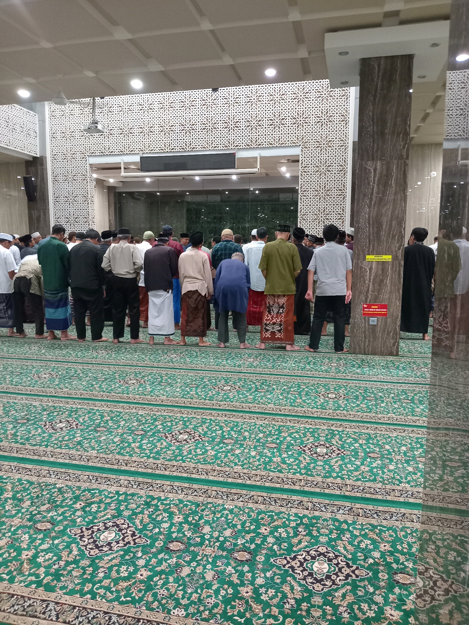A group of men stand in a mosque, facing forward and appearing to be engaged in prayer. They are dressed in traditional Islamic attire, including sarongs and peci hats. The mosque has a large, ornate carpet with a green base and intricate patterns. The walls are decorated with geometric designs, and large pillars support the ceiling. The overall impression is one of a religious gathering taking place in a well-maintained mosque in Randuagung, Indonesia.