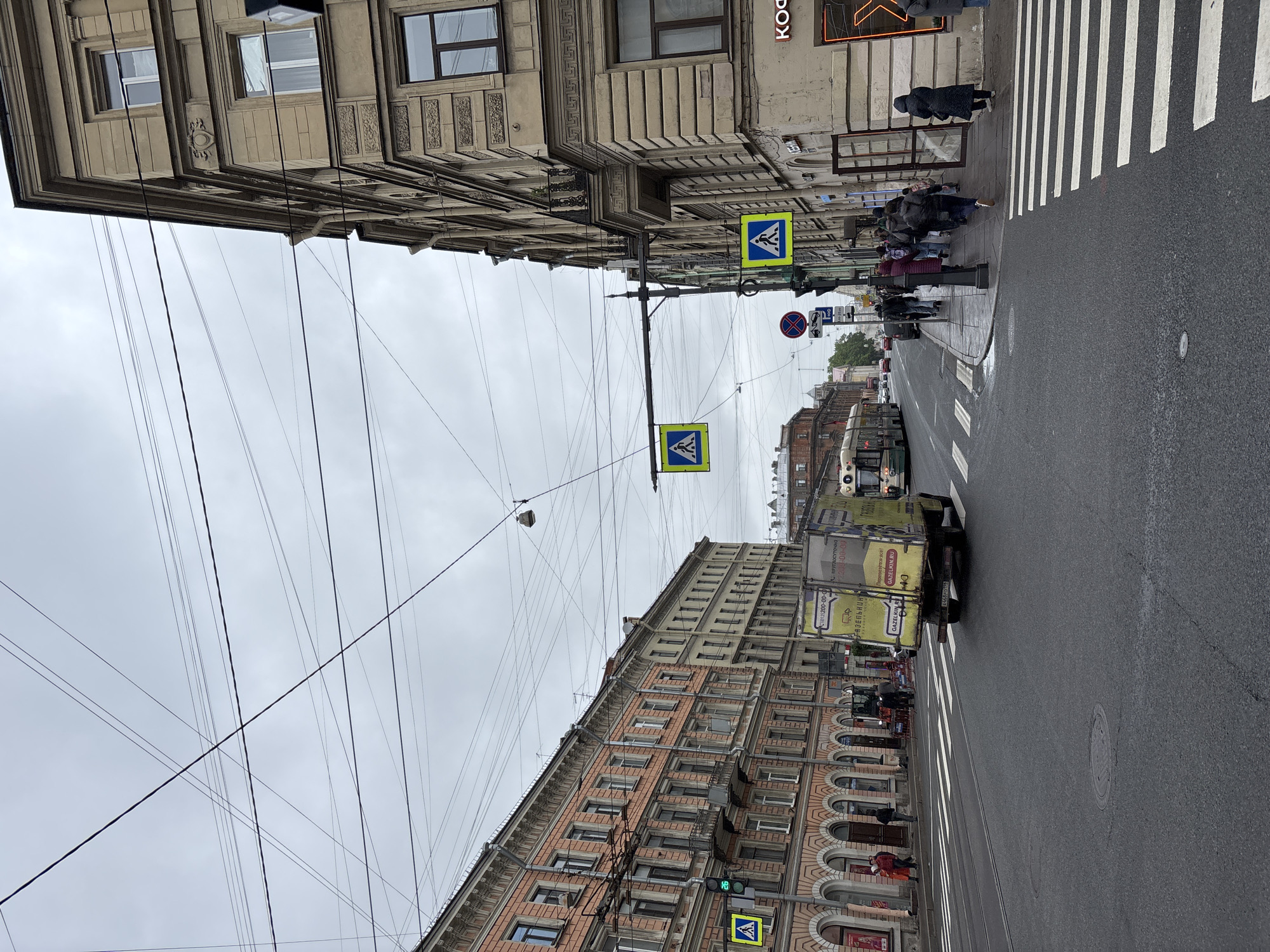 The image depicts a street scene in Saint Petersburg, Russia. The view is oriented along a straight, paved road with tram tracks embedded in its surface. Buildings with multiple stories line both sides of the street, showcasing a mix of architectural styles. The buildings on the left are characterized by a lighter color palette and ornate detailing, while the buildings on the right feature a combination of red brick and light-colored elements, along with arched windows. Overhead, a dense network of utility cables crisscrosses the sky, connecting to poles that hold pedestrian crossing signs.
A crosswalk spans the street, with pedestrians waiting on the sidewalks. A light-colored van is driving through the road. A bus stop is visible on the left side of the street, and street signs can be seen throughout the scene.
The environment appears overcast, with a gray sky dominating the upper portion of the frame. The overall scene conveys a sense of urban activity and historical architecture in Saint Petersburg.