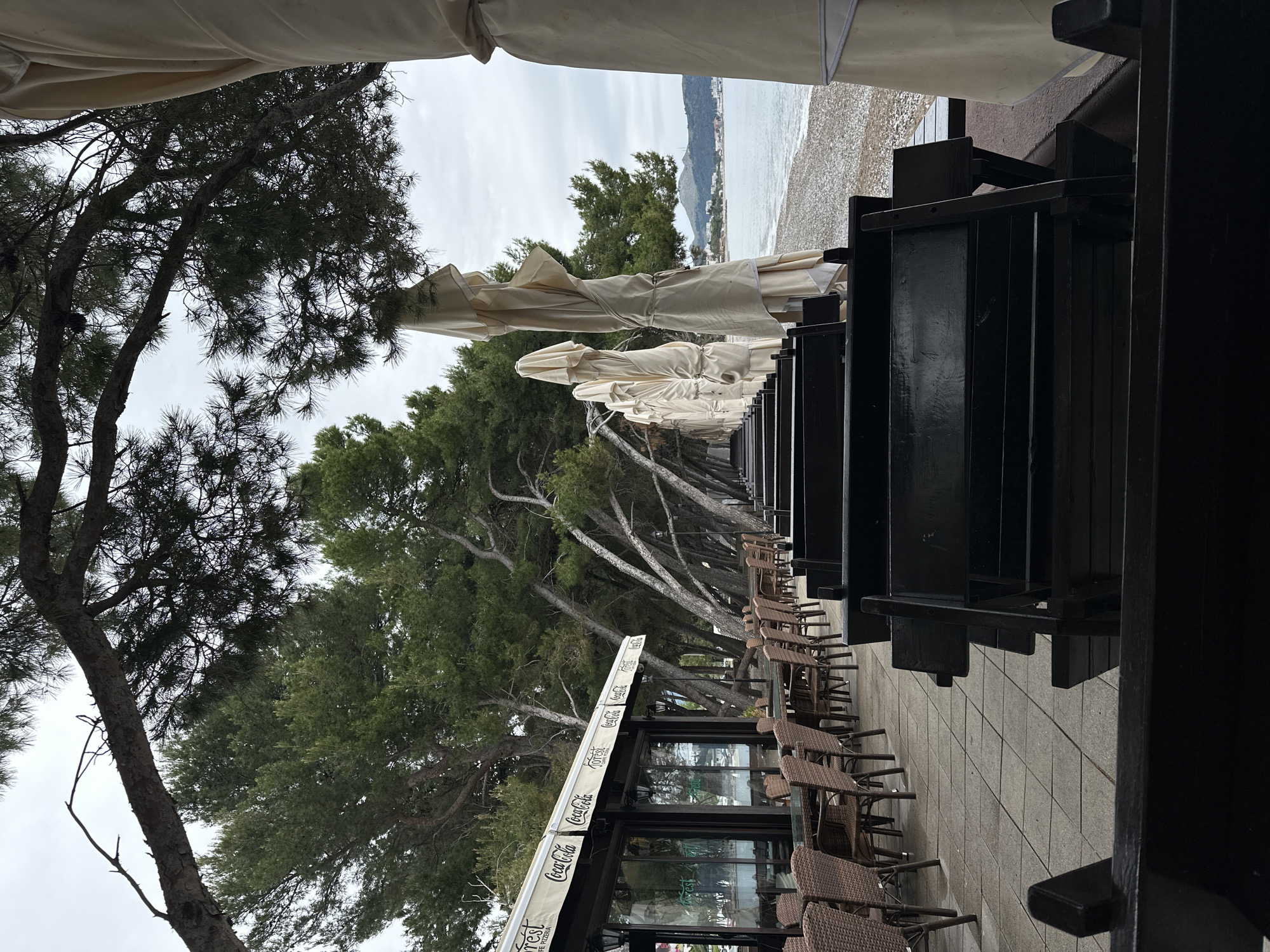 The image shows an outdoor restaurant or cafe in Šušanj, Montenegro, with a view toward the beach. A row of closed white umbrellas lines the outdoor seating area, which includes woven chairs and tables. The beach and ocean are visible in the background under an overcast sky. Pine trees are interspersed between the umbrellas and seating. A restaurant structure with a Coca-Cola branded awning is also visible. The furniture is dark and seemingly made of wood.