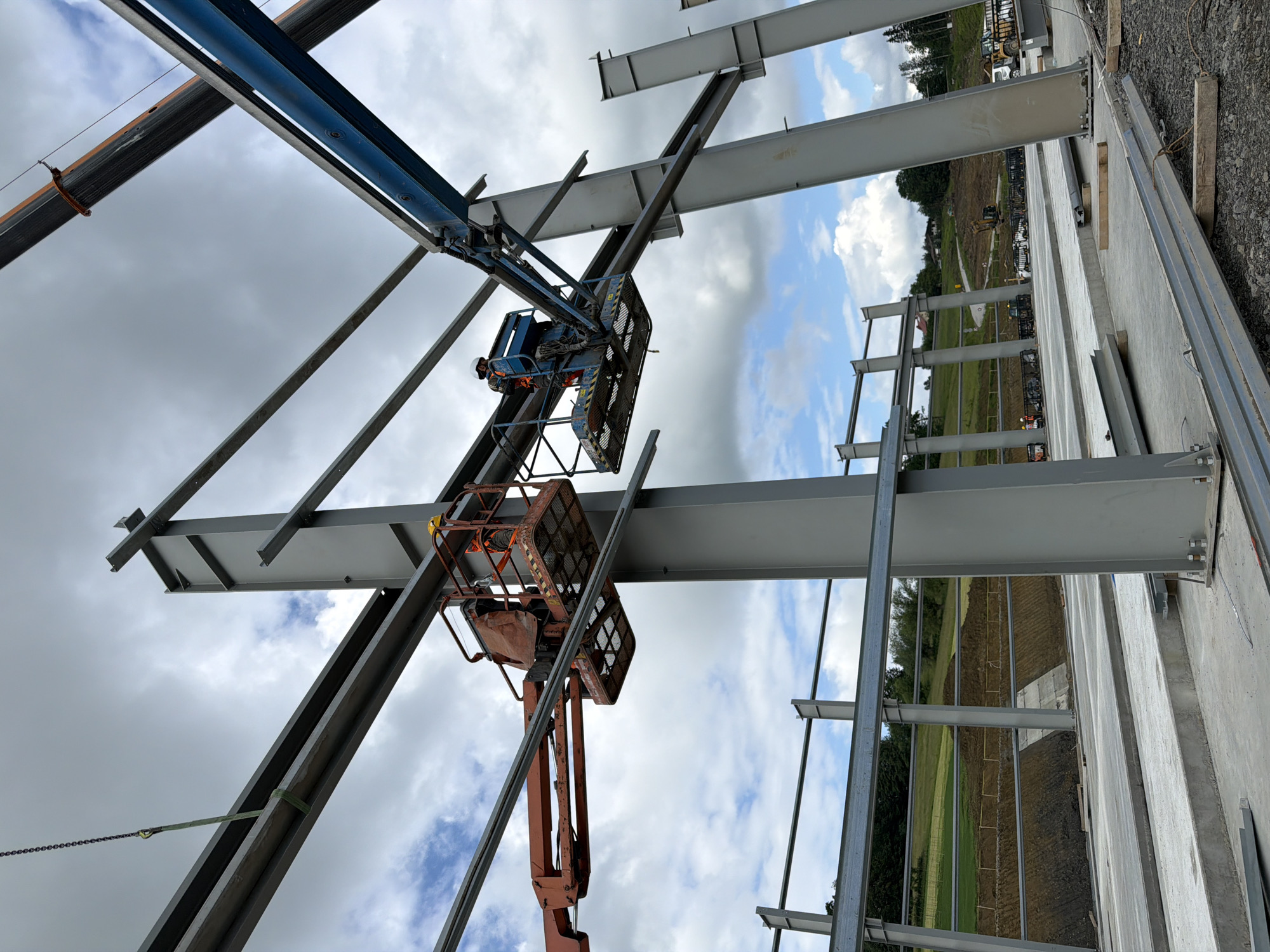 In Janowice, Poland, a construction site features a partially erected steel building. Two individuals, wearing hard hats and safety vests, are positioned in aerial work platforms. One platform, orange in color, holds a worker wearing orange and yellow safety gear. The other platform, blue, carries a worker in orange and grey safety attire. Both workers appear to be actively involved in the construction process, likely securing or positioning steel beams. The surrounding environment includes the large, grey steel framework of the building, concrete foundations, and raw earth grading in the background, indicative of ongoing construction activity. The sky above is cloudy.
