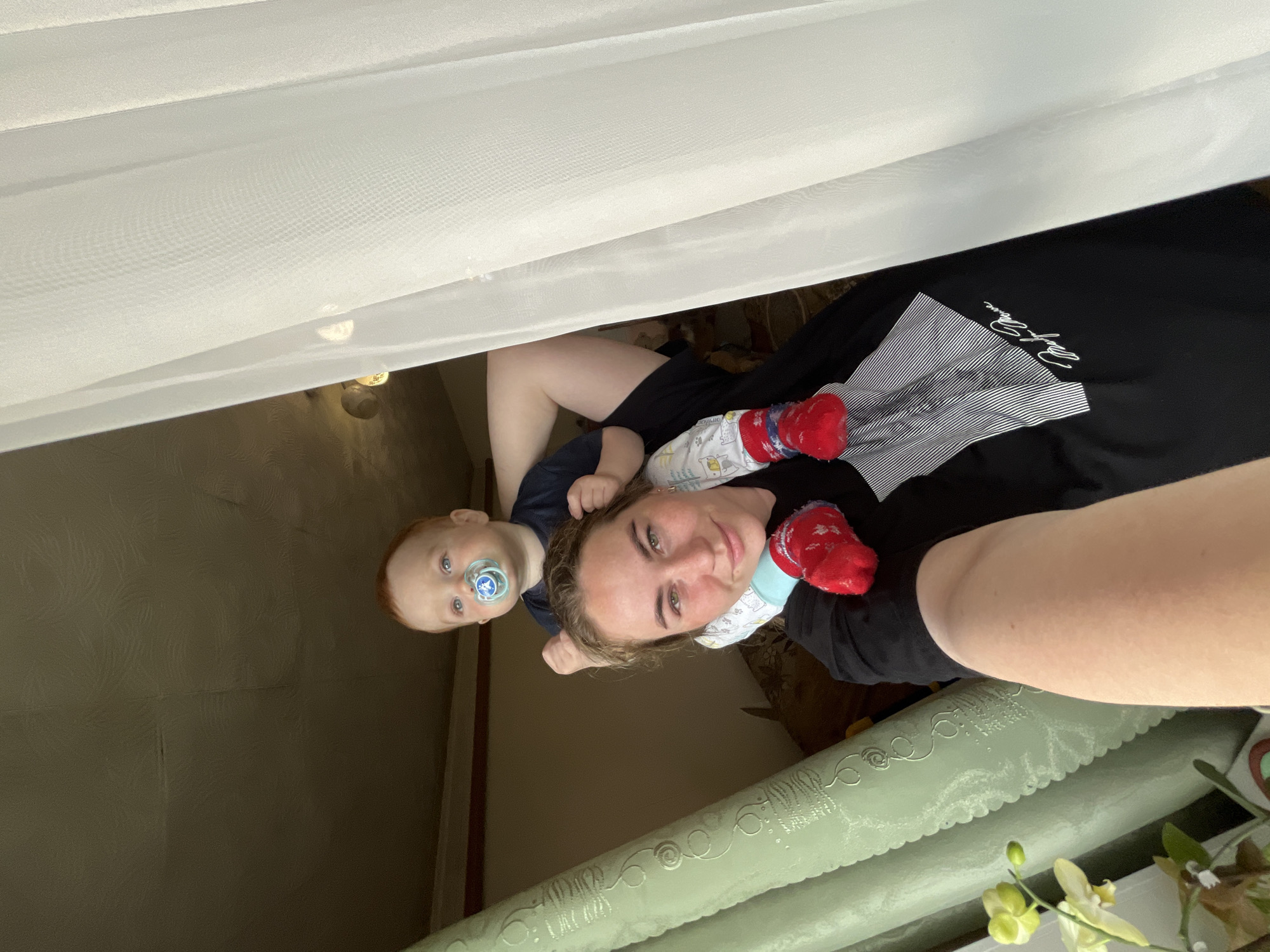The image depicts an adult female and a young child indoors, likely a residential dwelling, located in Сосново, Russia. The adult female, appearing to be in her 20s or 30s with light brown hair and green eyes, occupies the foreground, looking directly at the camera with a subtle smile. She wears a black short-sleeved t-shirt featuring a white striped geometric design and the cursive text "Marilyn Monroe". She is engaged in taking a selfie, with her right arm and hand holding the camera visible at the bottom of the frame.
A young child, approximately 1-2 years old with red hair and blue eyes, is seated on the woman's shoulders, looking slightly upwards and to the right of the frame. The child has a blue pacifier with a white animal design in its mouth and wears a dark blue top, white patterned leggings (depicting various animals), and red socks with white designs. The woman's left hand is visible, supporting the child's legs/feet.
The background features a white sheer curtain on the right side, indicating the presence of a window and a source of natural light. On the left, a light green, textured wall covering or curtain with a repeating swirl pattern is visible. The upper background shows a muted olive-green ceiling or wall with a textured surface, and a reddish-brown wooden beam runs horizontally across the upper middle section. A light fixture is dimly discernible behind the sheer curtain in the upper right background. In the bottom left, a light green orchid plant with two yellow-green blooms and one bud is partially visible on an unseen surface.