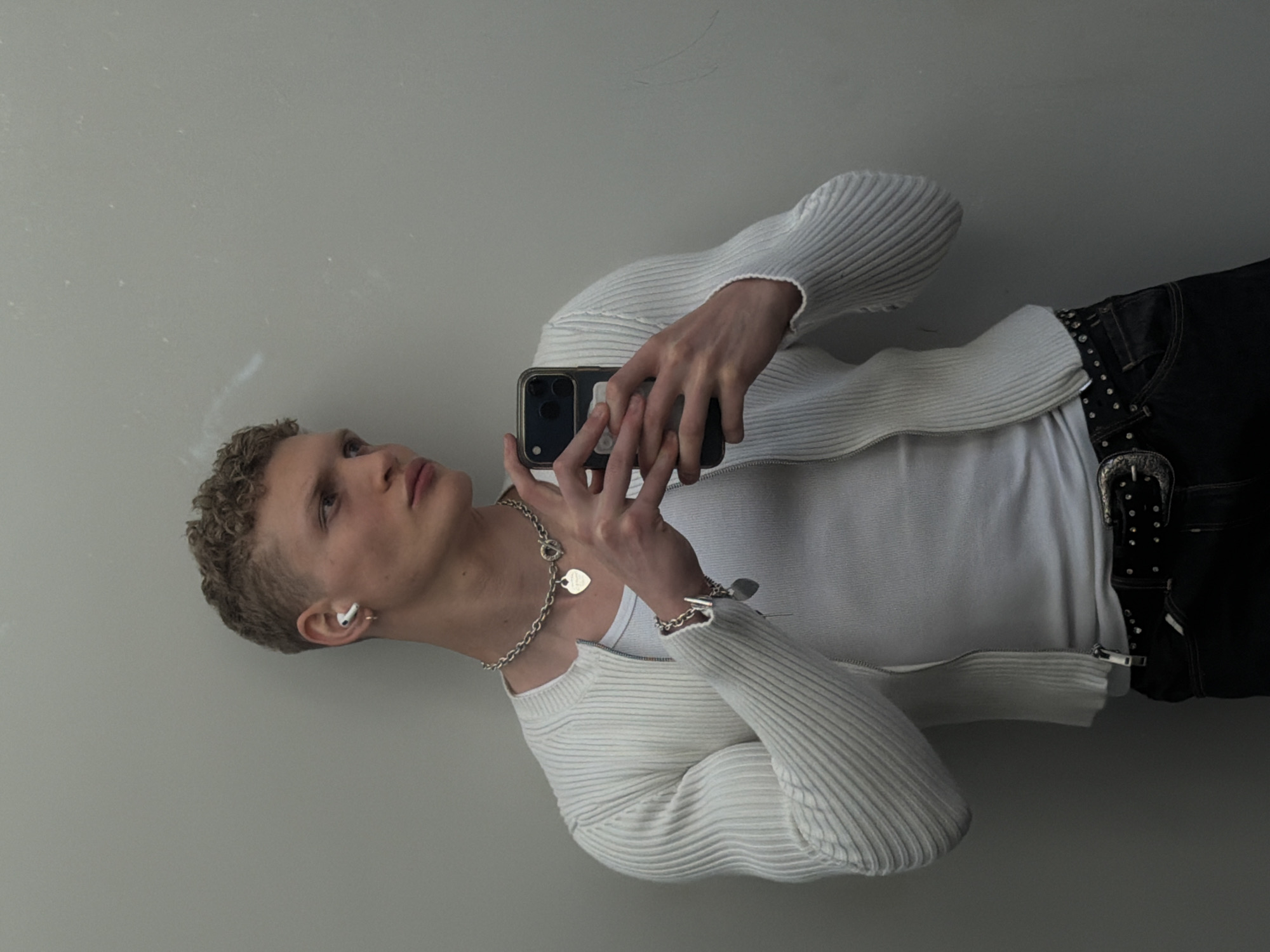 The image, rotated 90 degrees clockwise, depicts a young individual taking a selfie or video with a smartphone against a plain, light-colored wall. The person has fair skin, short, light-brown curly hair, and is looking slightly upwards and to the right of the frame with a neutral expression.
They are dressed in a white ribbed knit zippered cardigan worn open over a white t-shirt. Below, dark jeans are visible, cinched with a black studded belt featuring a large, ornate silver buckle with a western-style design. A silver chain necklace with two heart-shaped pendants (one larger, one smaller) is worn around their neck, and a matching silver bracelet adorns their left wrist. A white wireless earbud is noticeable in their left ear. Both hands grasp a dark-colored smartphone, which has at least two camera lenses visible on its back. The background is a simple, unadorned light grey or white wall, showing some minor marks or dust particles. There is no visible text in the image.