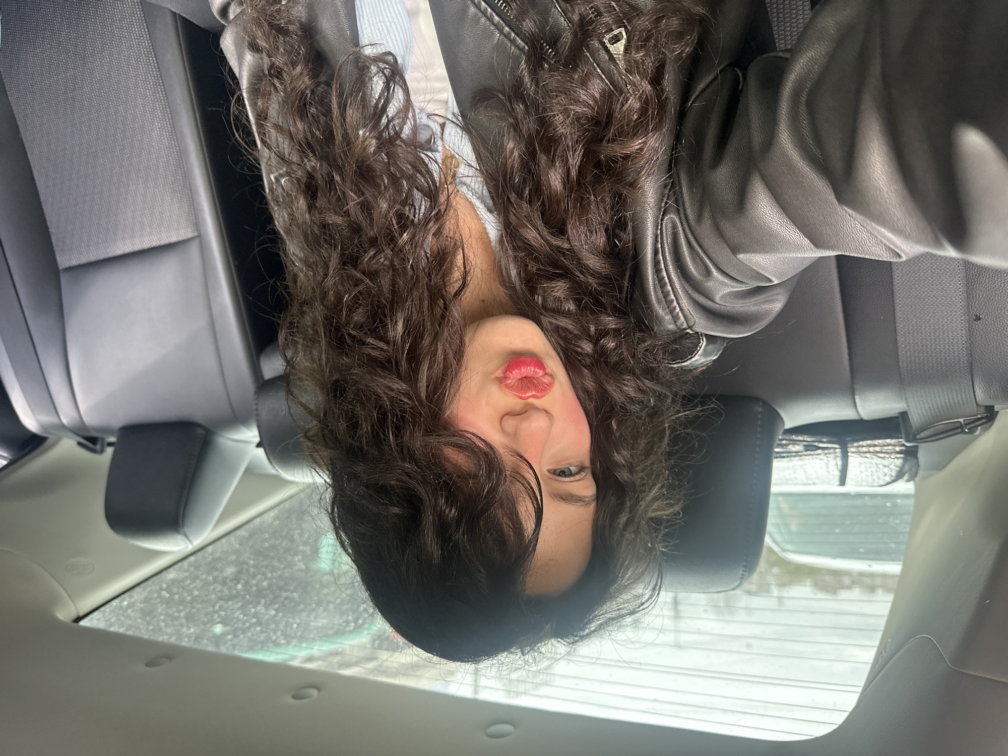 A woman is lying in the backseat of a car. She has long curly hair, bright red lipstick, and is wearing a black leather jacket. The car appears to be parked, as evidenced by the window showing a blurry exterior scene. The location cannot be determined from the image.