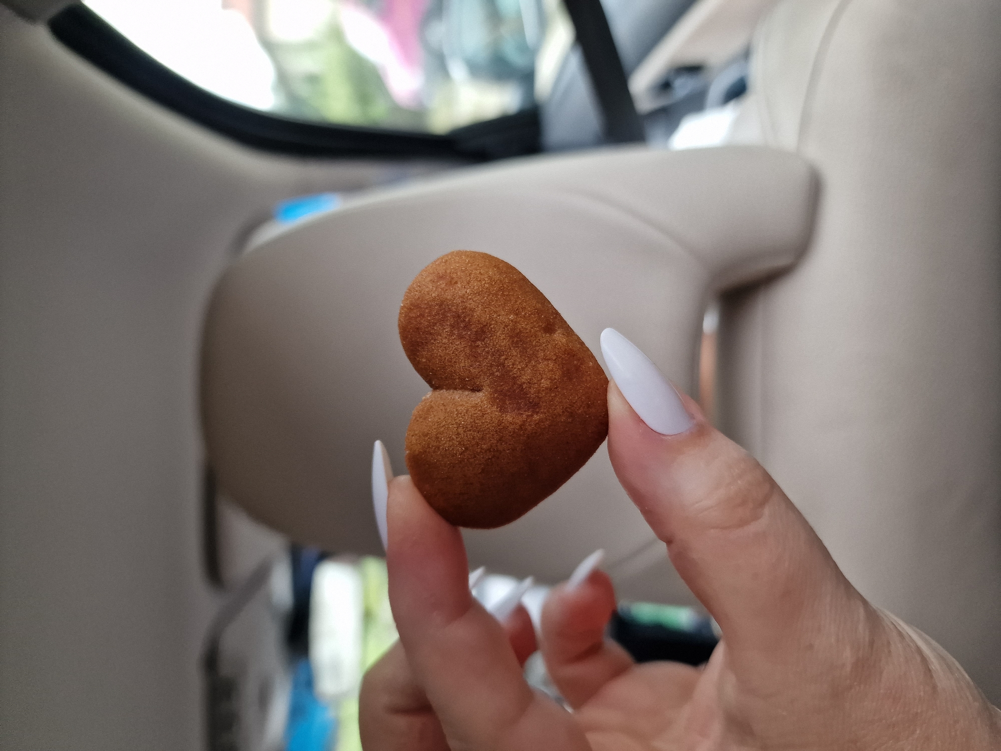 A close-up, eye-level shot shows a person's hand holding a small, heart-shaped cookie between their thumb and index finger. The cookie is a light brown color with a slightly textured surface. The hand has long, white, almond-shaped fingernails. The background is blurred but appears to be the interior of a vehicle, with a beige headrest visible. Through the car window, a blurry outdoor scene is visible, possibly a residential area with some greenery and a pink object that could be another vehicle. The photograph was taken in Troyan, Bulgaria.