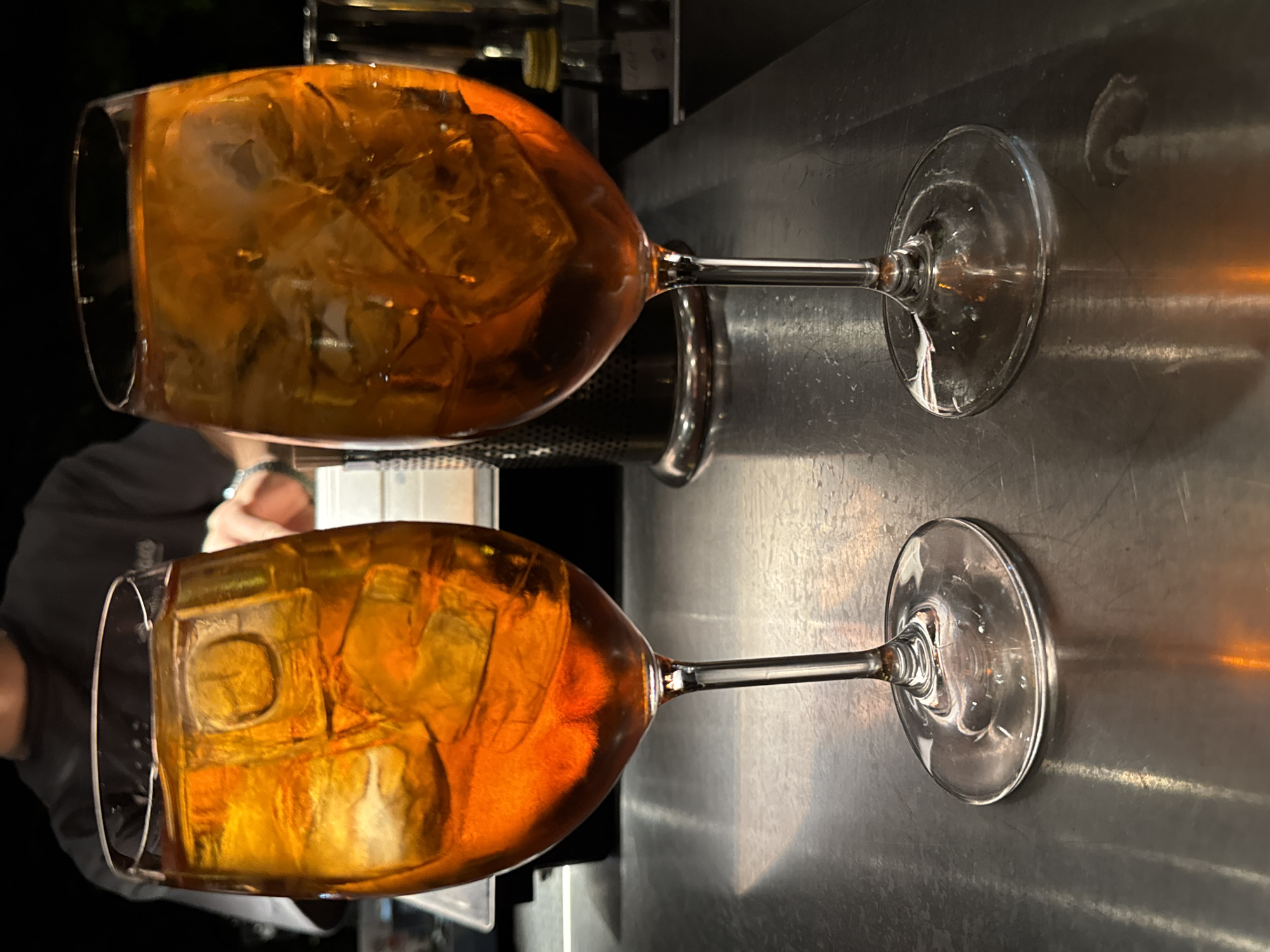 Two wine glasses filled with an orange liquid and ice are positioned on a metallic surface. The glasses are tall and have a slender stem. The liquid inside the glasses is translucent and reflects light, revealing the ice cubes within. In the background, a person wearing a dark t-shirt is partially visible, with their hands in motion, suggesting they might be a bartender or server. The environment appears to be a bar or restaurant setting. The scene is lit with artificial light, casting reflections on the metallic surface. No specific location in Yerevan, Armenia can be determined from the visual information provided.