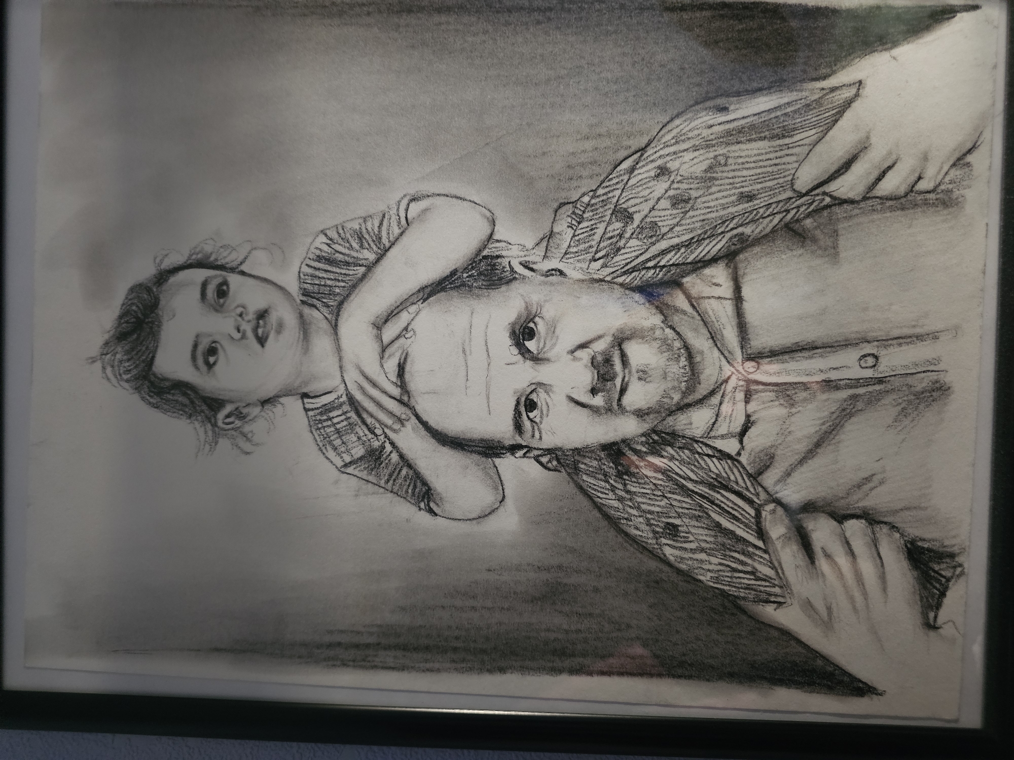 A framed charcoal drawing depicts a man lying down with a young child draped across his shoulders. The man looks directly at the viewer. The child is positioned on top of the man's head and upper chest. The man has a light complexion, short dark hair, and a visible beard. The child has fair skin, dark hair, and is wearing a patterned shirt. The man is wearing a button-down shirt with what appears to be another patterned garment underneath. The background is a blended grey charcoal texture. The image is contained within a dark frame. There is no indication of the location in Khimki, Russia.
