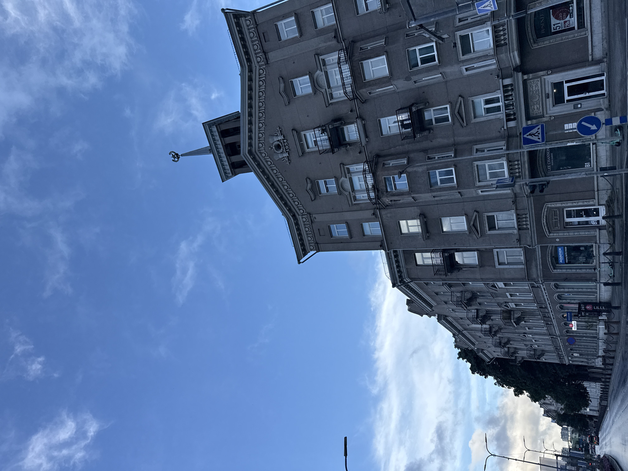The image depicts a street scene in Tallinn, Estonia, with a prominent, ornate grey building dominating the right side of the frame. The building features multiple stories, balconies with black railings, and decorative architectural details along the roofline and above some windows. A distinct tower with a spire and a decorative element resembling a crest or crown crowns the building's upper section. The year "1954" is visible above a central circular decorative element on the building's facade.
The street in front of the building appears to be wet, suggesting recent rain. Several streetlights and traffic signals are visible. On the lower level of the building, there are shop entrances with signage, including "LEE CHANNON," "B1," and "ROSE&E." There are also pedestrian crossing signs and a blue directional arrow sign on the street. On the left side of the image, more buildings and trees are visible, extending down the street. The sky above is blue with scattered white clouds. The perspective is from ground level, looking up at the building and the sky. There are no people clearly visible in the immediate foreground or interacting with the environment.