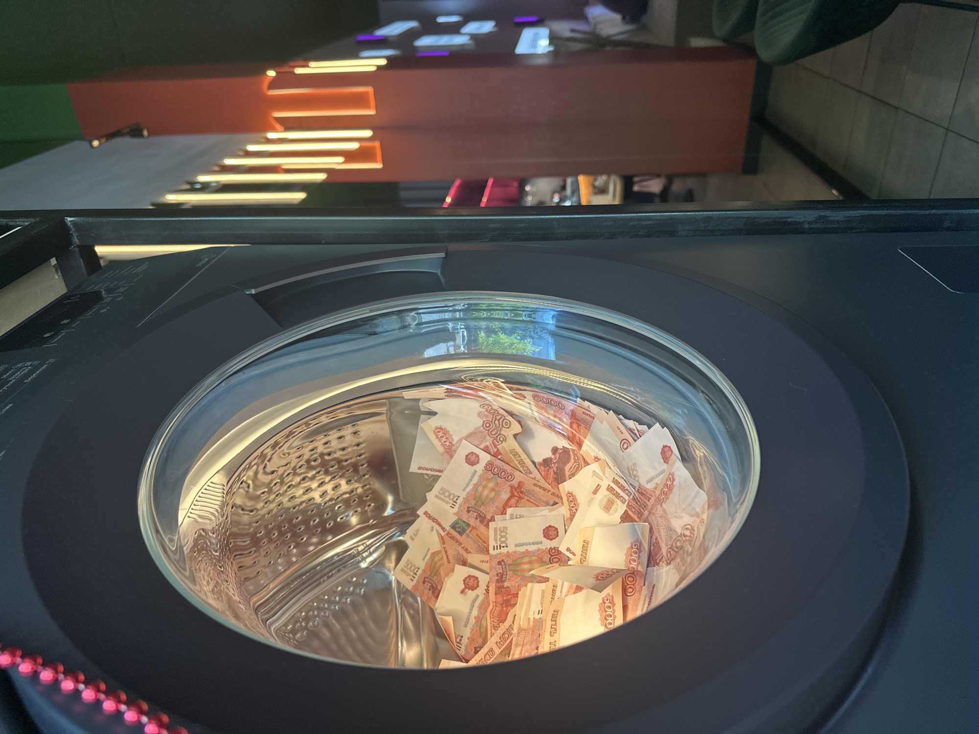 A high-angle shot shows a front-loading washing machine filled with Russian 5000 ruble banknotes. The washing machine is dark grey or black. Through the transparent door, the crumpled banknotes are visible. The interior of the machine appears to be stainless steel. The image's background is blurred, but it appears to be a modern interior space with lit architectural features. Based on the OCR text "BILLET BUY NOVOSIBIRSK", the photograph was likely taken in Novosibirsk, Russia.