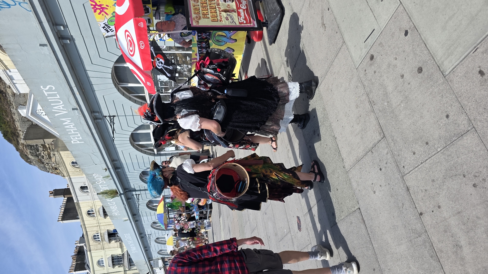 In St Leonards, United Kingdom, on a sunny day, a group of people dressed in pirate-themed attire are gathered on a paved walkway in front of a building with "PELHAM VAULTS" signage. One individual, facing away from the camera, wears a black top and a voluminous, colorful skirt, with a large drum strapped to their back. Another person, also in a pirate costume with a black hat adorned with red feathers, stands to the right, holding a drink. A third person, similarly dressed as a pirate, is partially visible. Behind them, a stall with a red and white umbrella displays a "MIX IT UP!" sign and what appears to be ice cream. Further in the background, other stalls and people are visible, as well as a multi-story building with arched windows. The ground is paved with large rectangular stones, and shadows are cast by the people and objects. The overall scene suggests a festive event or reenactment.