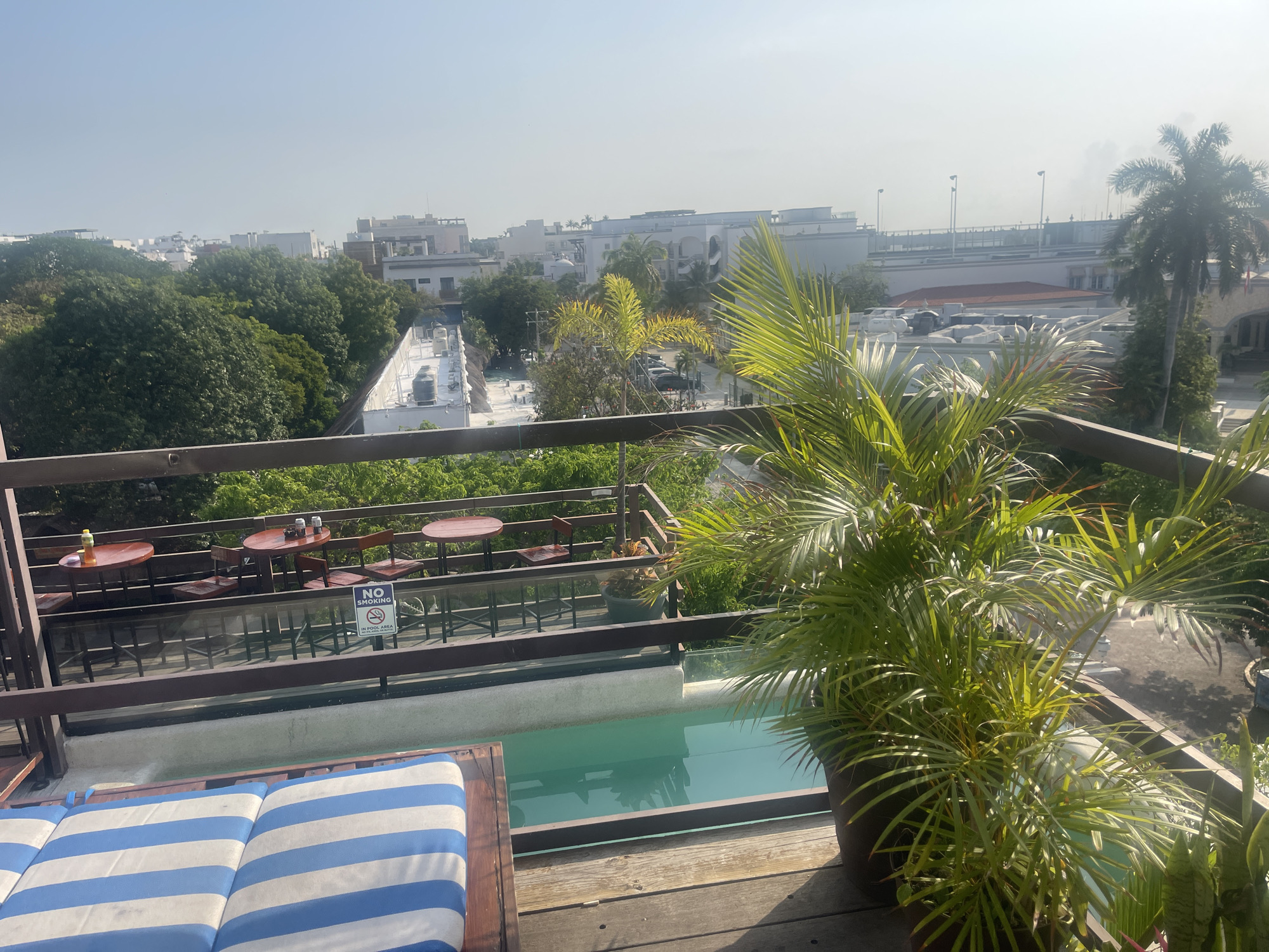 The image depicts a rooftop scene in Playa del Carmen, Mexico. In the foreground is a wooden deck with a blue and white striped lounge chair. A small pool with turquoise water is visible behind the lounge chair. To the right of the pool is a large potted palm. A railing separates the foreground from a dining area with small, round tables and chairs. A "No Smoking In Pool Area" sign is attached to the railing. Beyond the dining area, the cityscape of Playa del Carmen is visible, including buildings, trees, and a highway overpass. The sky is hazy and bright.