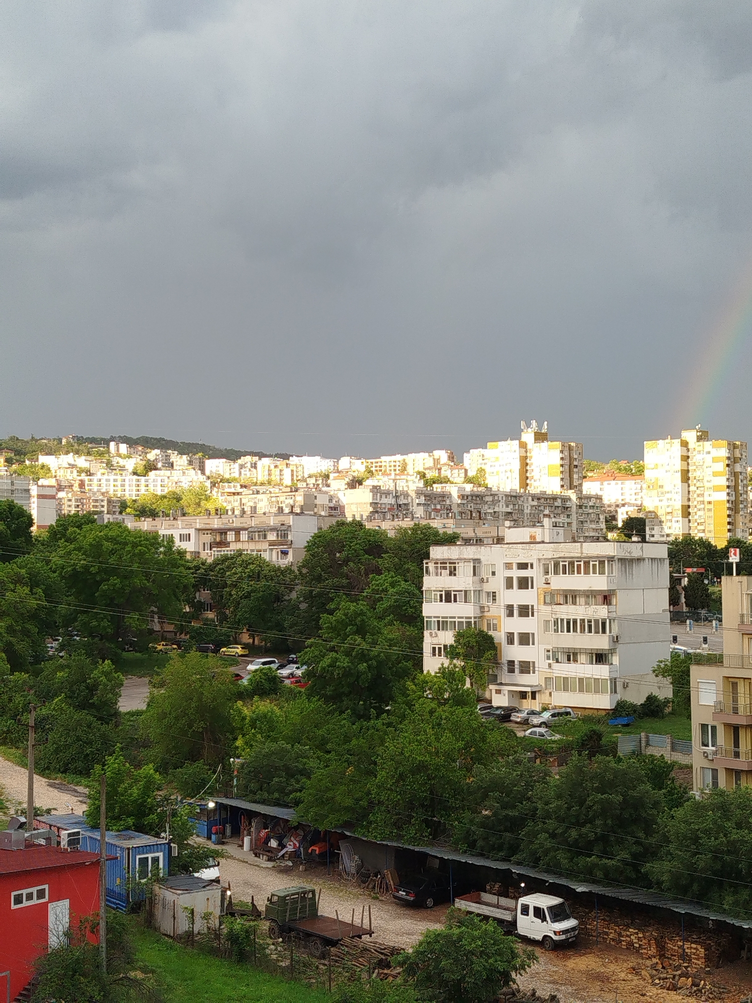 A view of Varna, Bulgaria reveals a cityscape interspersed with abundant greenery. Apartment buildings in light hues, including white and yellow, are seen under a sky that shifts from overcast with a prominent rainbow on the right to areas brightened by sunlight. At the bottom of the image are buildings and structures of varying sizes and colors, including a red building, a blue shipping container, and an open shed or workshop where people seem to be active. Assorted vehicles are parked nearby, including trucks and vans. The buildings and the vegetation suggest the image was taken during late Spring or early Summer.