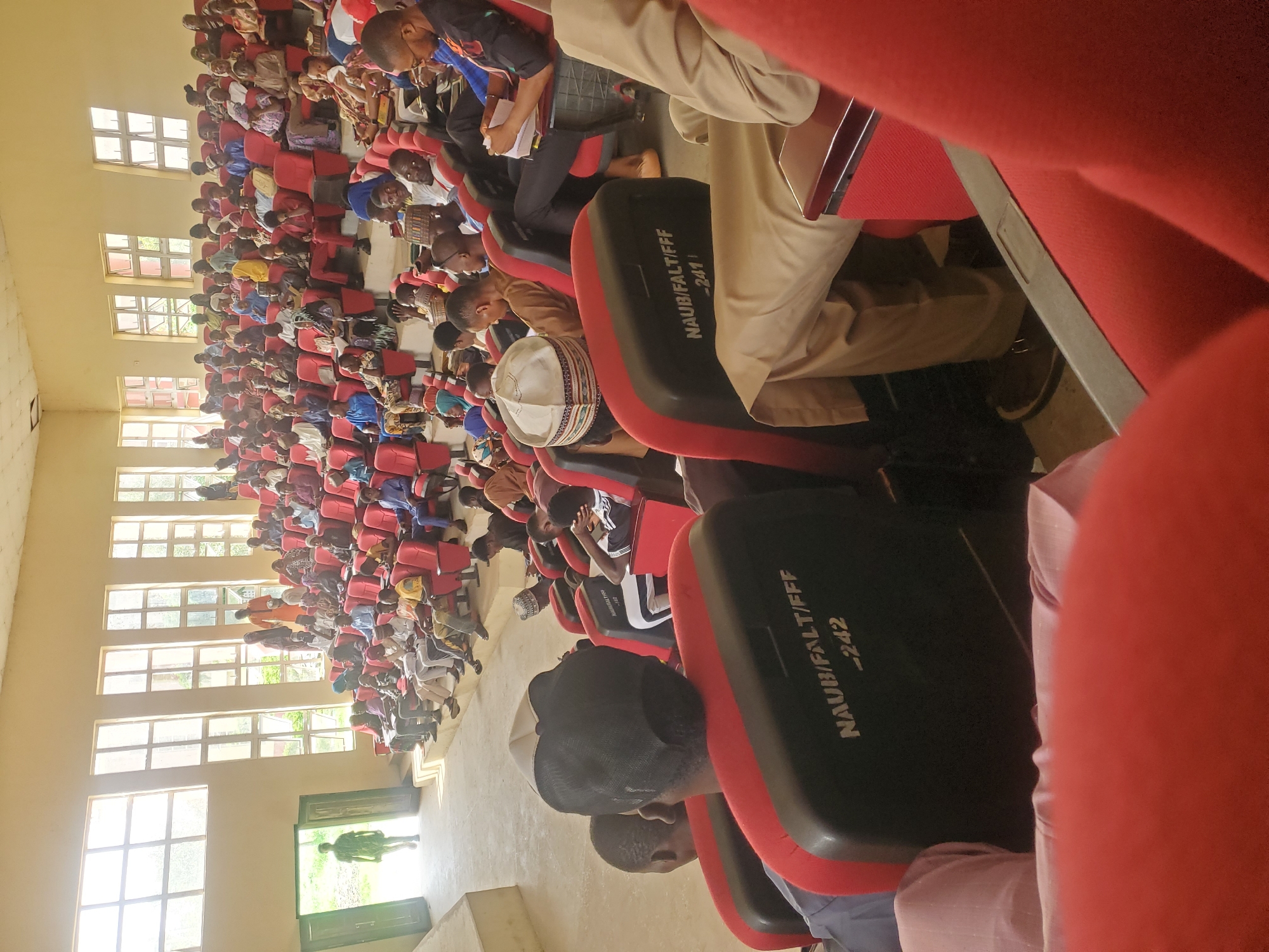 The image shows a medium shot of an audience seated in an auditorium, facing towards a stage area. The auditorium has red tiered seating with black backrests. The backrests are labeled with "NAUB/FALT/FFF" and numbers like "-242". Sunlight streams in from large windows on the left side of the room, illuminating the front of the auditorium. A person in a green shirt is walking through an open doorway on the far left, entering the auditorium. The audience members are diverse in age and attire, with many wearing traditional African clothing and head coverings. Some are looking towards the front, while others are engaged with their phones or conversing. The overall atmosphere appears to be that of a gathering or lecture. The city and specific location within the city are unknown.