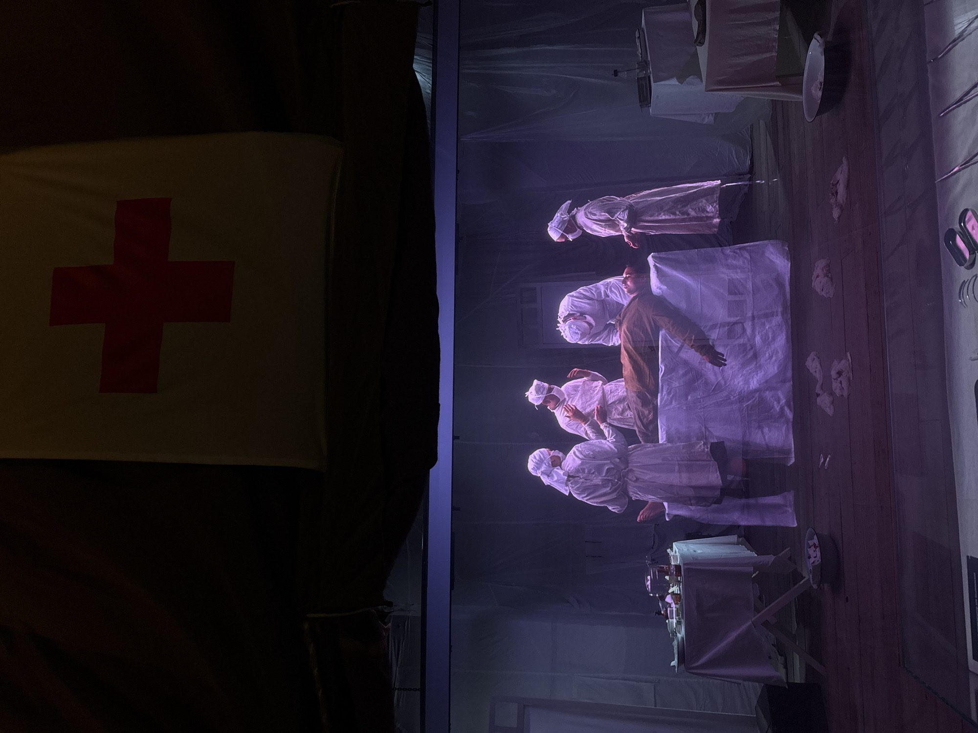 The image depicts a staged scene of a medical operation, possibly in a historical context, set within the city of Zelenaya Roshcha, Russia. In the center, a figure lies on an operating table, covered by a white sheet. Four individuals in medical attire, wearing white gowns and surgical caps, are positioned around the table, appearing to be surgeons or nurses. One individual is leaning over the operating table, possibly performing the procedure. To the left and right of the operating table are smaller tables covered with white cloths, holding various medical instruments and containers. Above the scene, a yellow banner with a red cross is prominently displayed, suggesting a connection to a medical organization or historical period. The background is dimly lit, with draped fabric or curtains creating a theatrical atmosphere. The floor appears to be made of wooden planks. The overall impression is a reenactment or display of a surgical procedure.