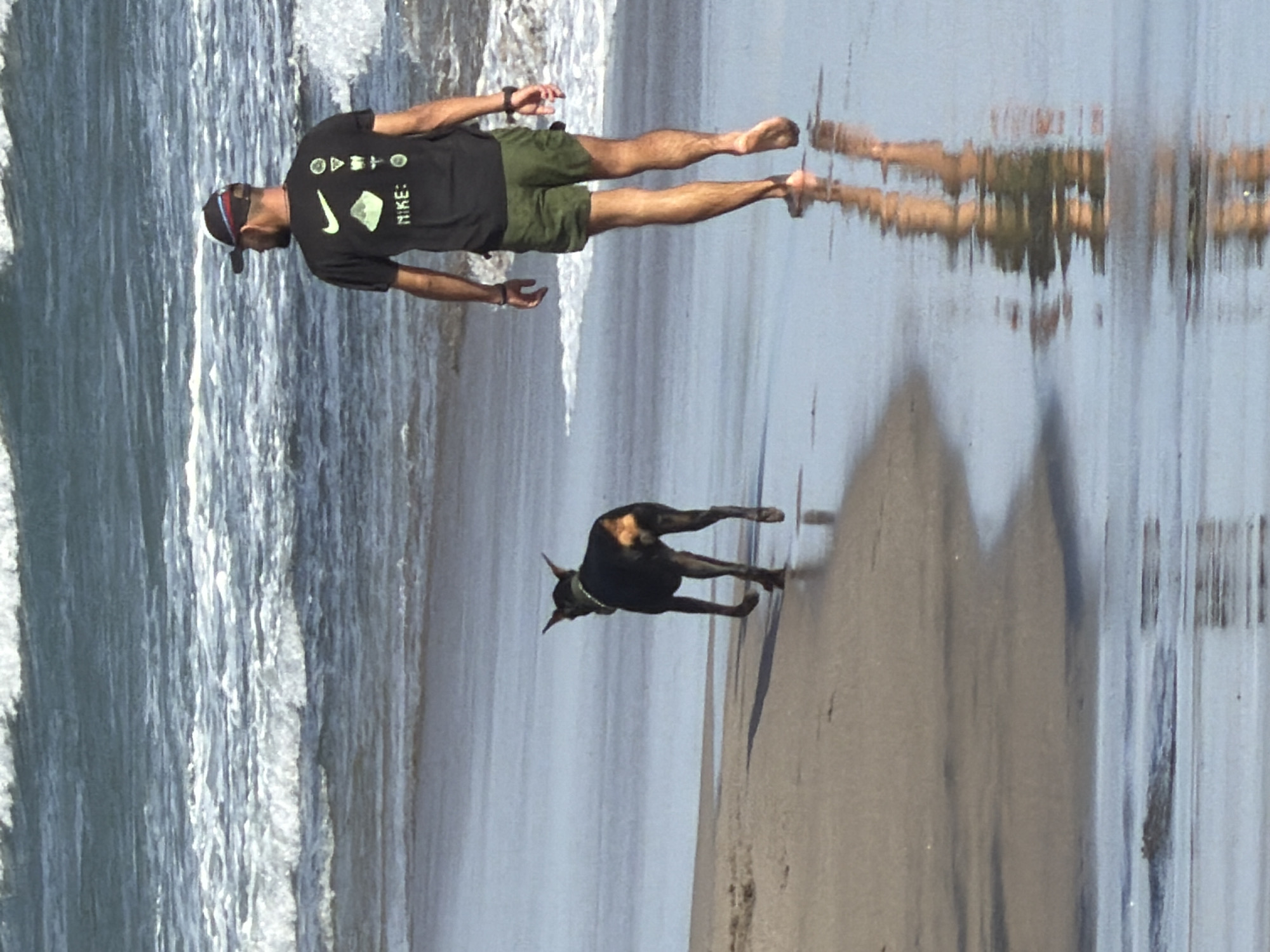 A man and a dog walk along the wet sand of a beach. The man is wearing a black t-shirt with a Nike logo and other graphics, green shorts, and a baseball cap. He is walking away from the camera towards the ocean, with his left arm extended slightly. The dog, a black Doberman with tan markings on its face and hindquarters, is walking in front of the man, also away from the camera. The dog is wearing a collar. The ocean is in the background, with waves breaking on the shore. The sand is wet and reflects the sky and the figures. The location is the beach in North Kuta, Bali, Indonesia.