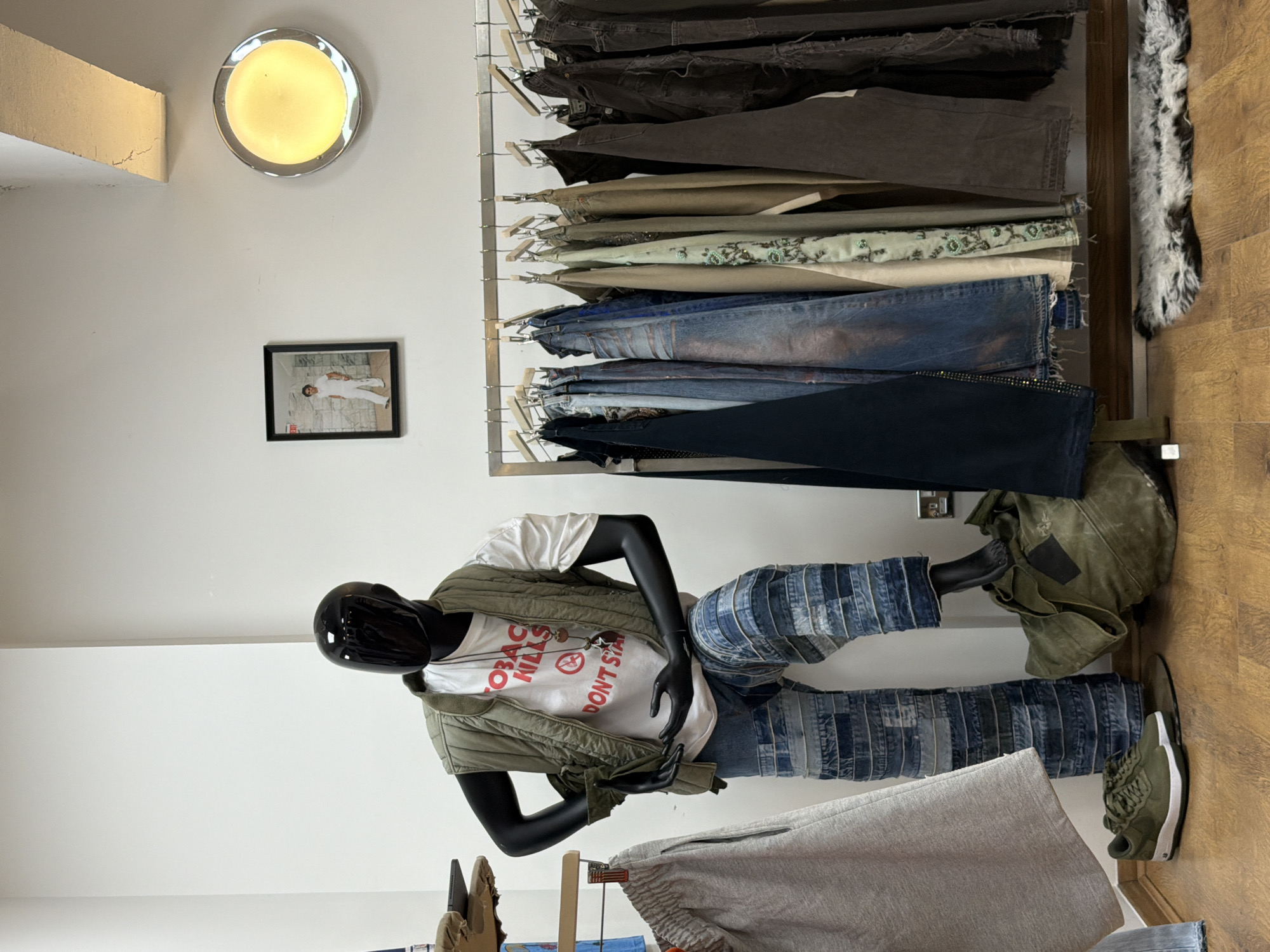 The image captures an indoor retail space. On a light-colored wall, a circular illuminated mirror and a framed picture hang. A clothing rack displays an array of garments, primarily trousers or jeans in various shades and materials. In front of the rack, a mannequin is dressed in a white t-shirt with a print, a vest, and patchwork jeans. Next to the mannequin, an olive green bag and sneakers are placed on the wooden floor. Additional clothing is visible near the bottom of the frame. The location, city, and country are not discernible from the image.