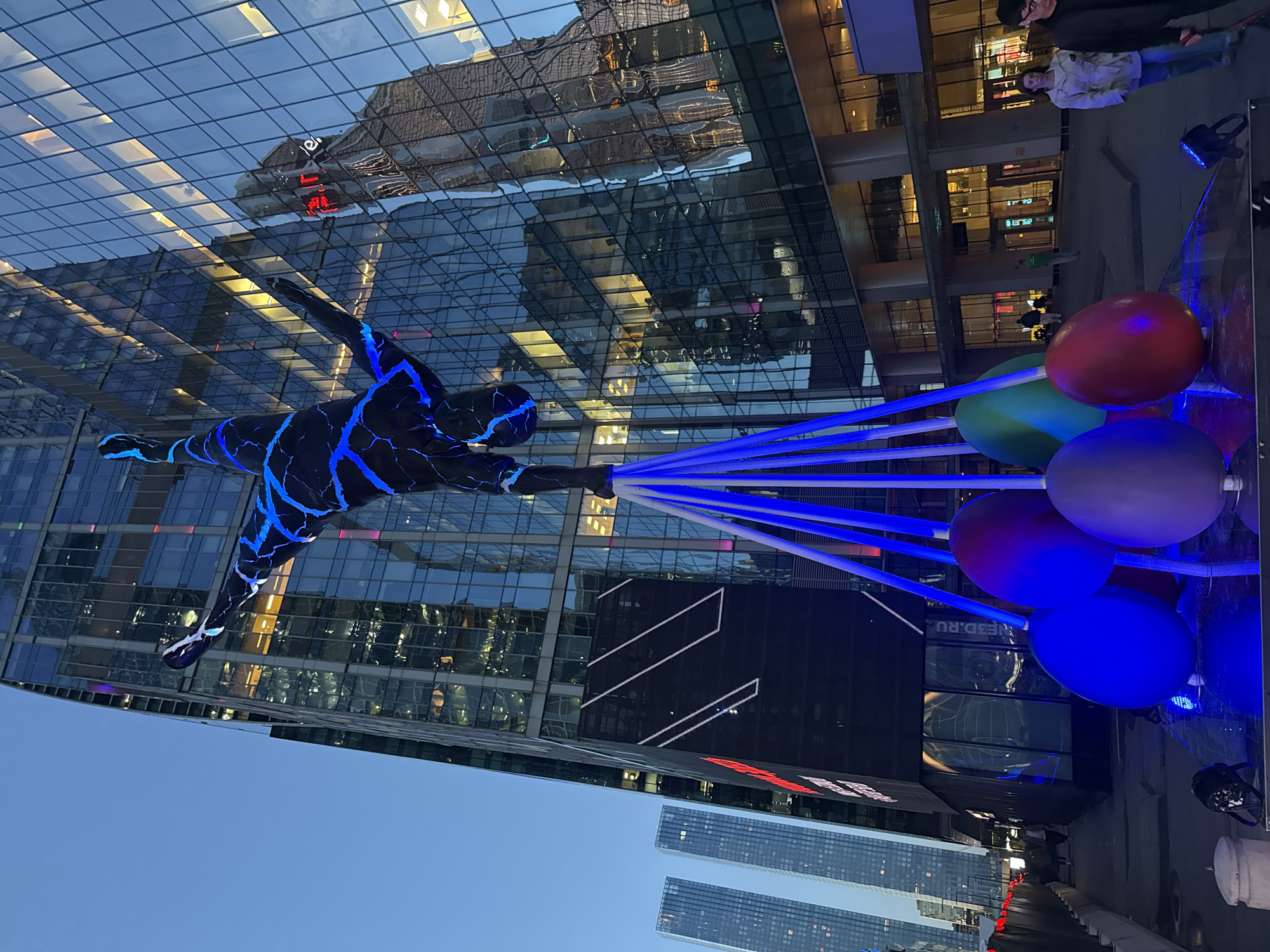 This media file depicts an outdoor nighttime scene in Moscow, Russia, featuring a striking art installation. The primary subject is a large, illuminated sculpture of a human figure, outlined with bright blue, crackling light patterns against a dark body, appearing to be in motion. This figure is tethered to a cluster of large, colorful balloons – red, green, and blue – by a series of bright blue beams of light.
The setting is a modern urban environment, characterized by towering glass-fronted buildings. The reflective surfaces of these buildings capture and distort the surrounding lights and architecture, creating a complex visual tapestry. In the foreground, a few people are visible, seemingly observing the installation. One couple stands near the balloons, their faces partially illuminated by the artwork. The time of day is clearly night, indicated by the artificial lights and the dark sky. The weather appears to be clear.
One notable detail is the text "NE3D.RU" visible on a black screen or surface within one of the buildings. The overall mood conveyed is one of wonder and technological artistry, set against the backdrop of a vibrant city at night. The scale of the sculpture and the dynamic lighting suggest a public art display or a festive event.