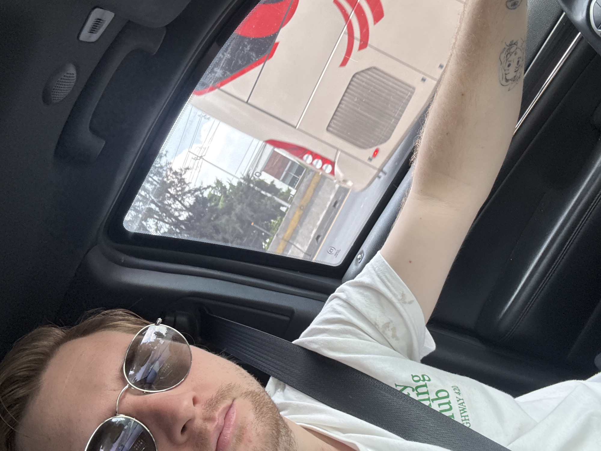 A male with blond hair and a light beard, wearing sunglasses, a white t-shirt with green text that reads "HIGHWAY 420", and a black seatbelt, is driving a vehicle. He is looking at the camera. Through the car window, a beige and red bus is visible on a road. The surrounding environment appears to be an urban or suburban area with trees and buildings. The scene is captured from inside the car, suggesting a personal photo taken while traveling. The image is dated and located in Cuautitlán Izcalli, Mexico.