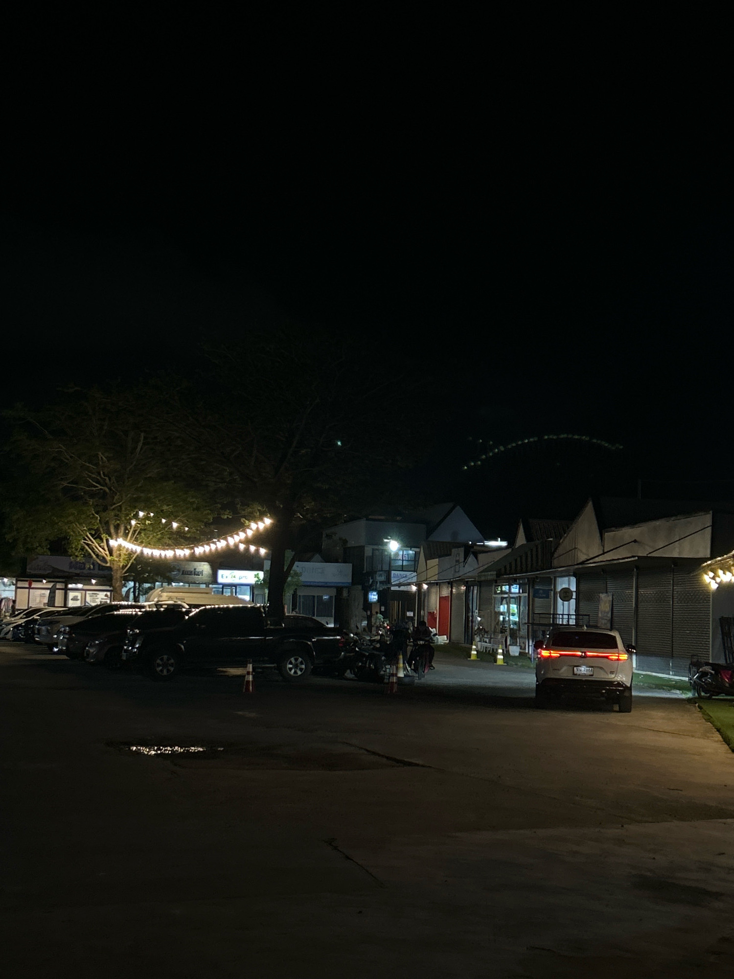 A nighttime scene depicts a parking lot in Ratsada, Thailand, illuminated by strings of white fairy lights draped between trees and on the fronts of buildings. In the foreground, the asphalt ground shows a wet patch. Several vehicles are parked, including a black pickup truck with a traffic cone beside its rear passenger wheel. To the right, a white SUV with its brake lights illuminated faces away from the camera. In the background, a row of shops with distinct rooflines line the street. A few individuals are visible, including one on a motorcycle and another walking. The sky is dark, with faint lights suggesting distant structures or a light show in the upper right quadrant.