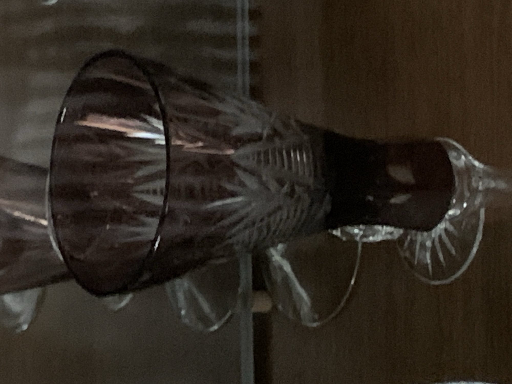 The image shows a close-up, eye-level view of a stemmed glass with a dark purple hue and intricate etched patterns on its bowl. The glass appears to be made of crystal and is positioned on a dark wooden surface. The background is blurred, suggesting a shallow depth of field, and consists of indistinct shapes and lighting that do not provide specific environmental cues. There is no indication of a specific location within Tashkent, Uzbekistan, or any people, actions, or interactions.