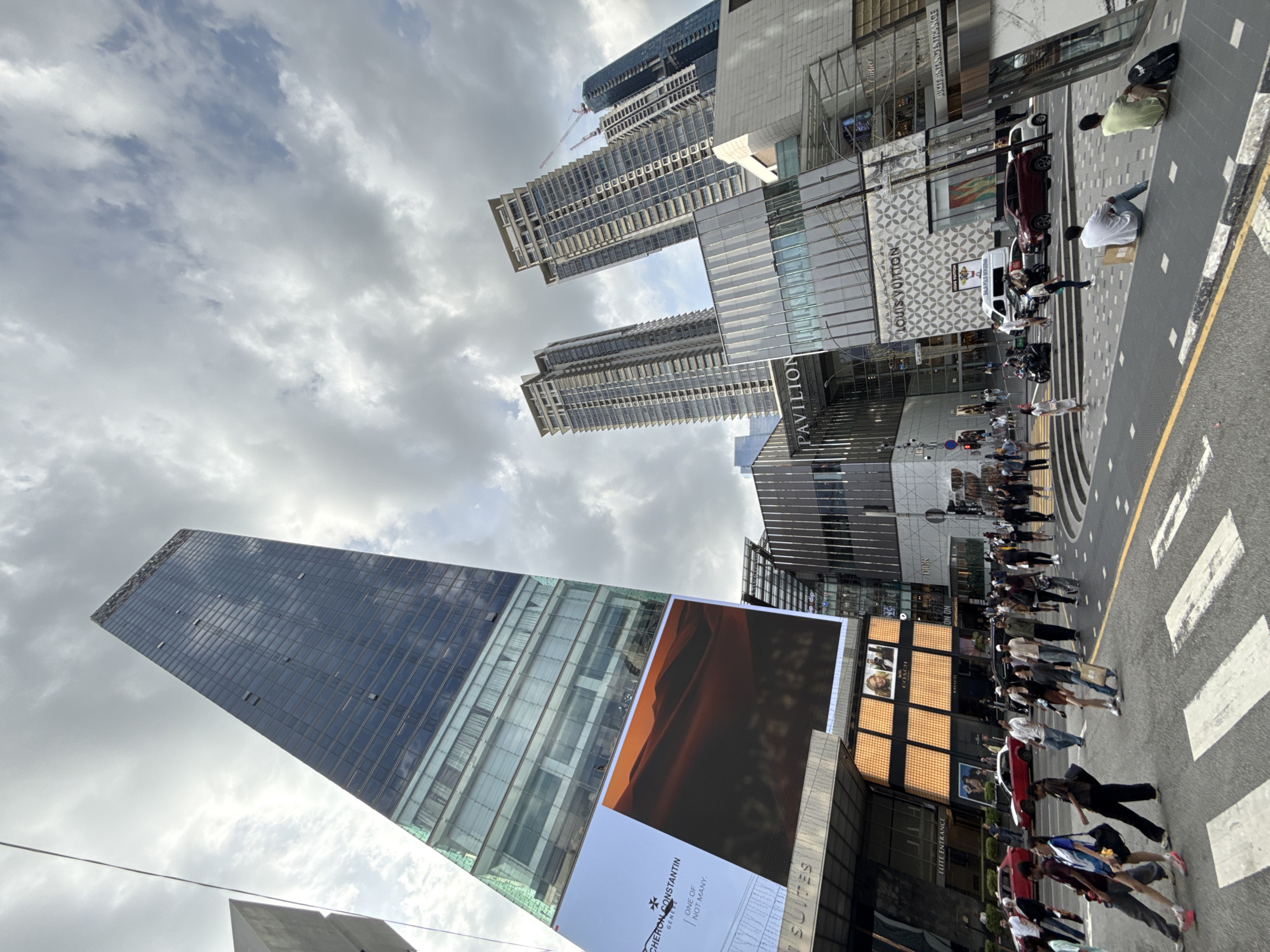 The image, taken in Kuala Lumpur, Malaysia, is angled upwards, capturing a blend of urban architecture and street activity. On the left, a tall, modern skyscraper dominates the frame, partly covered with a large advertising screen featuring a Vacheron Constantin ad. Adjacent to it are other buildings, one displaying "Pavilion" signage, and various stores, including Louis Vuitton. A street scene is visible on the right, where a pedestrian crossing is actively used by numerous people. Further back, there are vehicles and more storefronts. The sky is overcast with a mix of grey and white clouds.