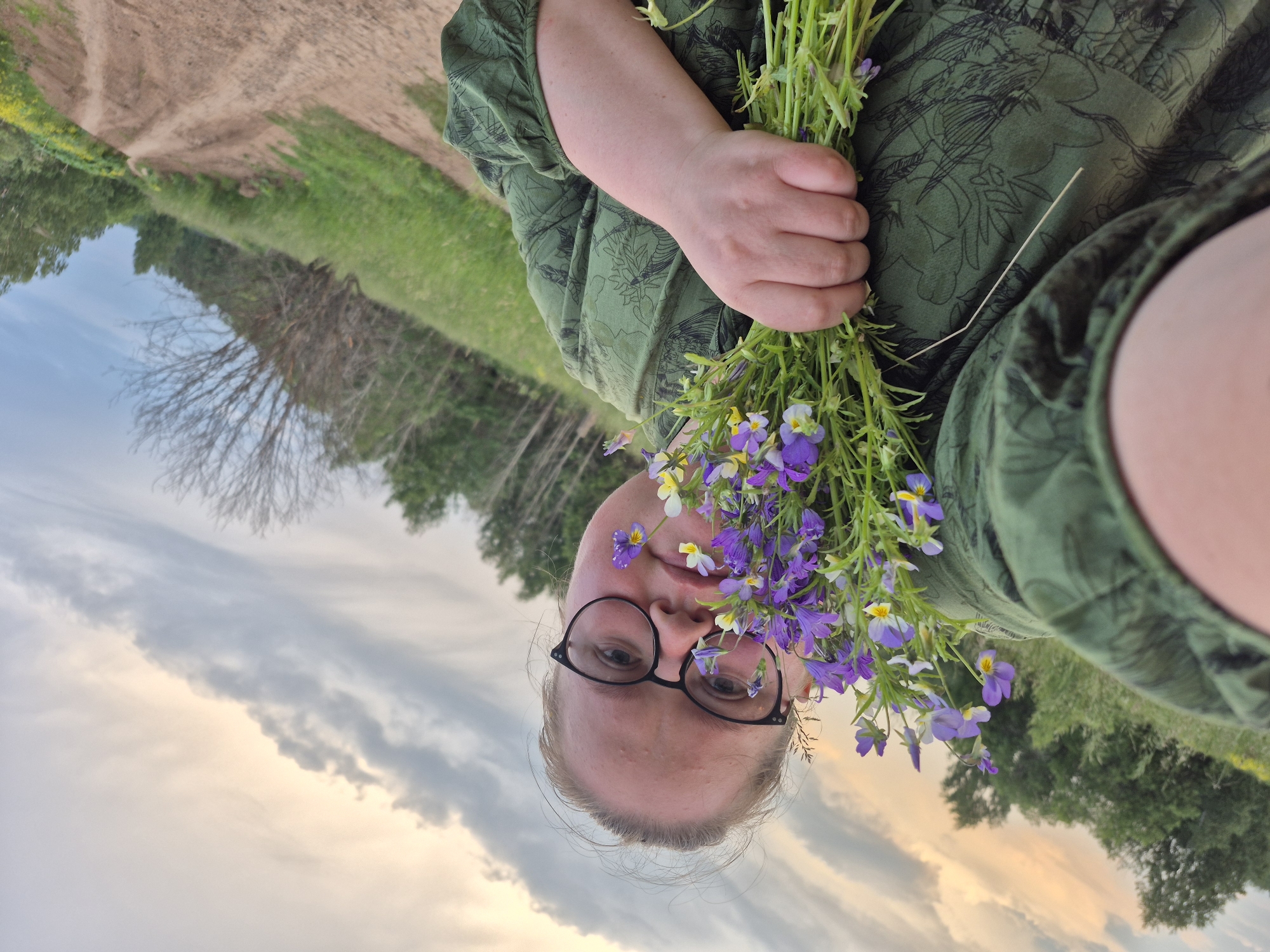 In a selfie taken from a low angle, a person with blonde hair and glasses is holding a bouquet of small purple and yellow wildflowers. The person is wearing a green dress with a floral pattern. They are outdoors, with a dirt road and grassy fields visible in the background. Trees and a cloudy sky with hints of sunset are also present. The overall scene suggests a rural or natural setting, possibly in or around Doinaya, Russia, given the context.