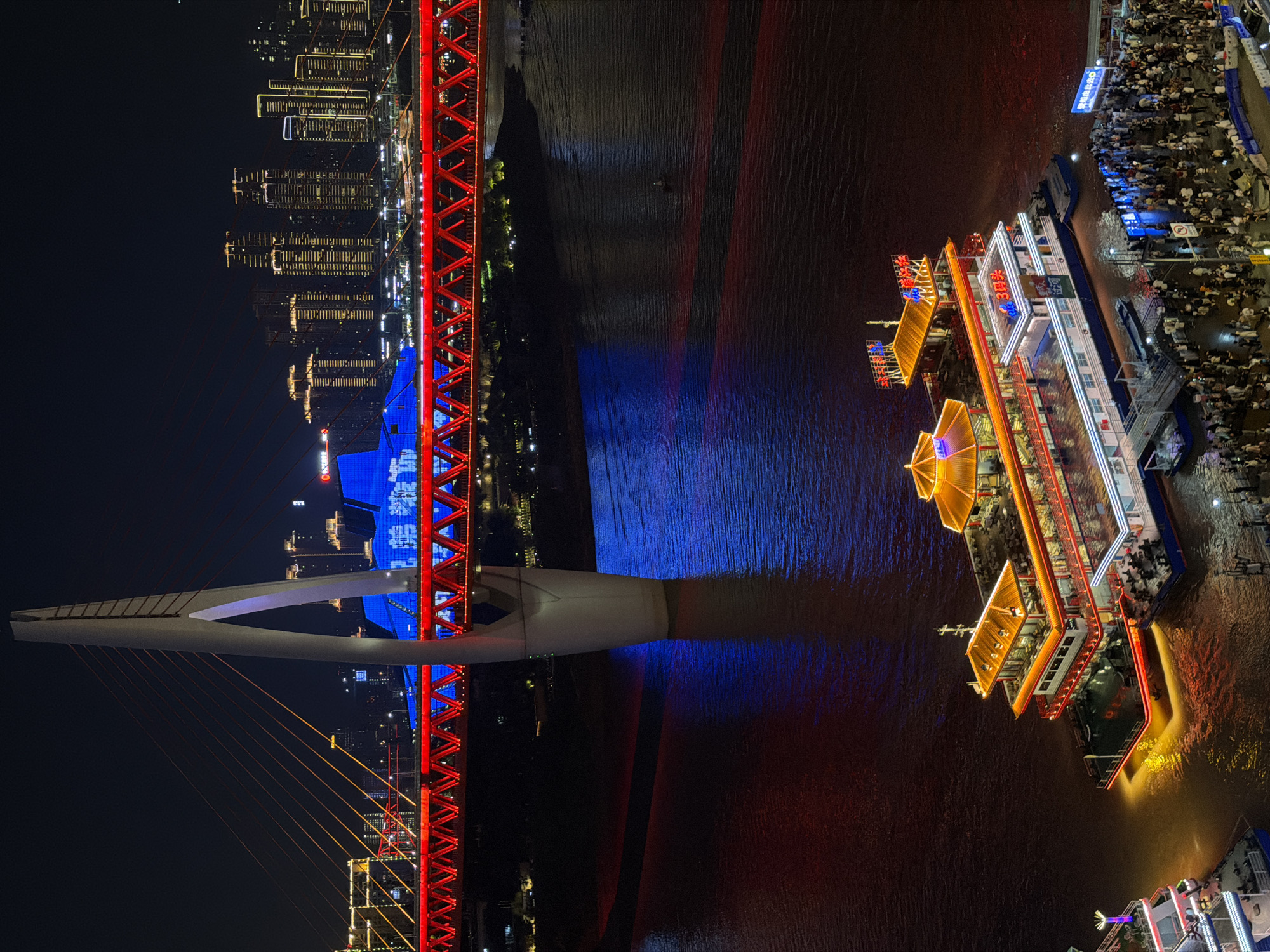 The image captures a night view of the Yuzhong District in Chongqing, China. In the foreground, a large, multi-story pleasure boat, illuminated with warm orange and red lights, is docked at a pier. Numerous people are gathered on the pier and boarding the boat. Adjacent to it, another smaller boat is partially visible.
Dominating the mid-ground is a vibrantly lit suspension bridge, with its red lattice structure and supporting tower illuminated against the dark night sky. The bridge spans a wide body of water, likely a river, where reflections of the bridge and city lights shimmer. In the background, a dense skyline of modern high-rise buildings, also aglow with lights, stretches across the horizon, signifying the urban development of the area. The overall scene is a dynamic interplay of light and architecture, showcasing the lively nighttime atmosphere of the city.
