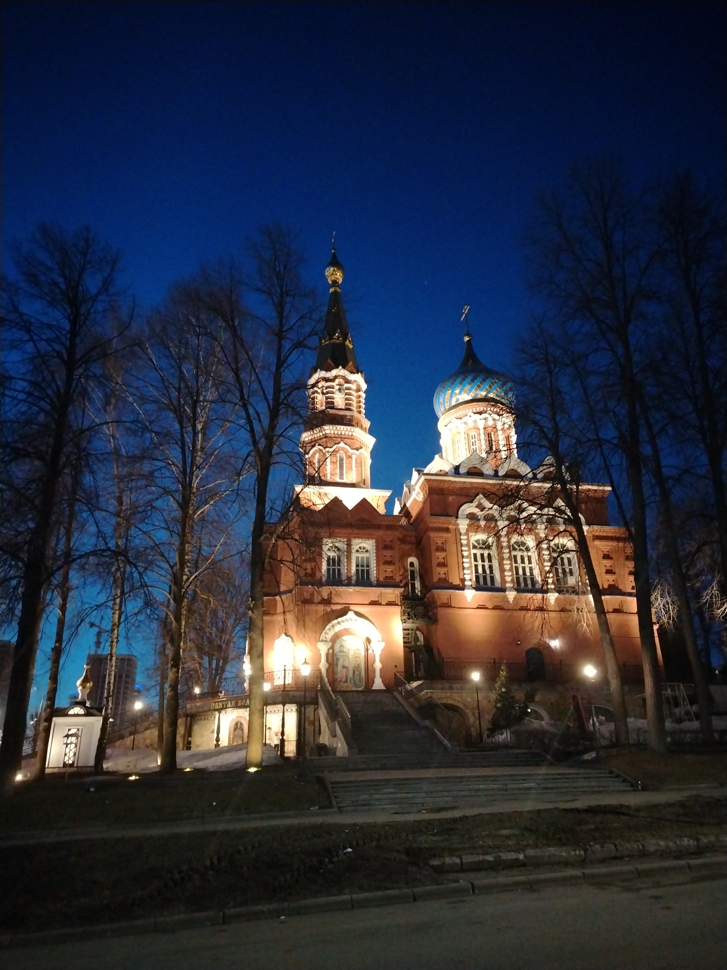 Captured at night, the image portrays a lit-up red brick Russian Orthodox church in Izhevsk, Russia. The church's architecture features an ornate tower on the left and a blue and white striped dome on the right. Lit windows and wall lights illuminate the building. Bare trees frame the church on both sides. Steps lead up to the church entrance. The foreground features a street and some grass. A small building is visible to the left of the church. The sky is dark blue.