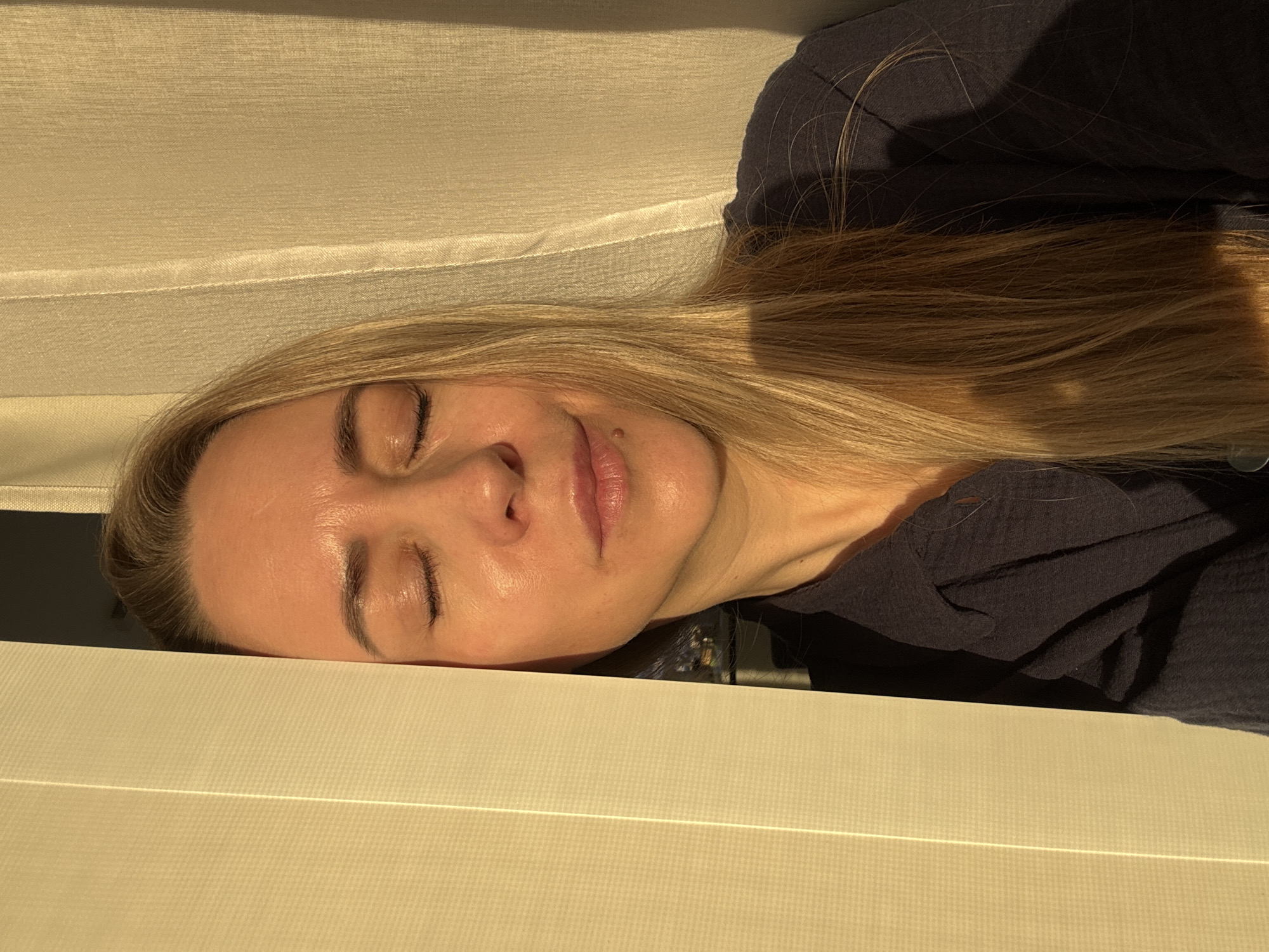 The image, taken in Chaikovsky, Russia, presents a close-up of a person, likely a woman, resting or sleeping peacefully indoors. Her head is tilted to the left, with her eyes closed and a subtle, serene expression on her face, including a slight smile. She has fair skin and long, straight, light blonde hair that flows around her head and shoulder. A small, light-colored mole is visible just above her upper lip on the left side of her face.
She is wearing a dark blue or charcoal grey garment, possibly a shirt or top, visible around her neck and shoulder. The setting appears to be an interior space, with the woman lying on or against a light beige or cream-colored, textured fabric surface, possibly a pillow, bed, or upholstered furniture. This surface has distinct horizontal lines or seams.
The scene is bathed in warm, golden sunlight, indicating either early morning or late afternoon. The direct light brightly illuminates her face, creating a soft glow and subtle highlights on her skin, while casting some shadows on her hair and the dark fabric of her clothing. The overall mood conveyed is one of tranquility and repose. There is no visible text in the image.