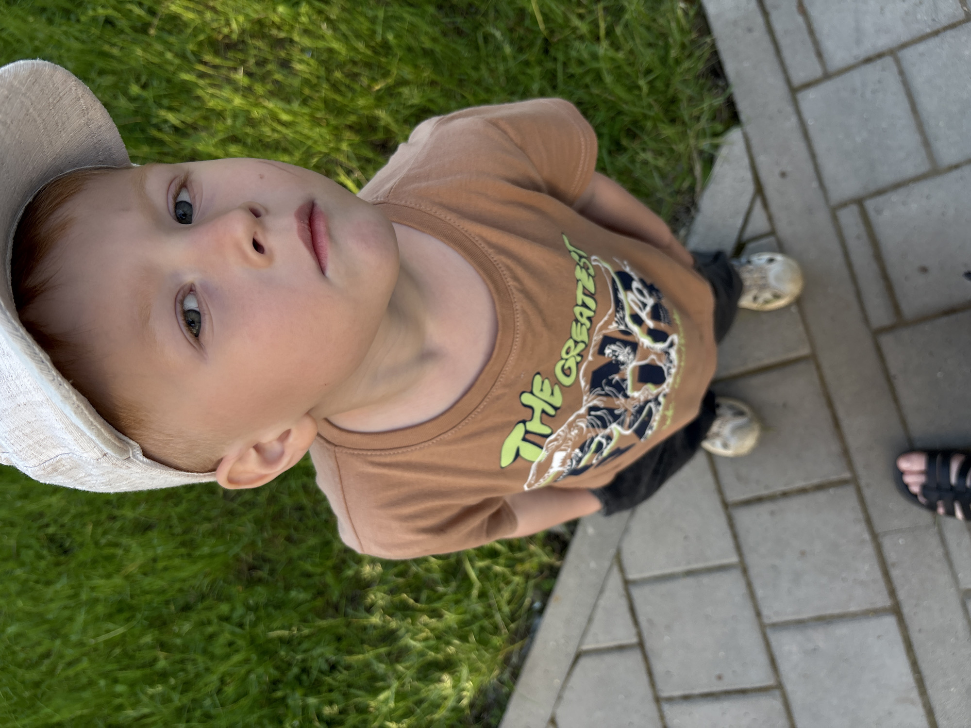 A young boy wearing a light-colored cap and a brown t-shirt with a graphic print stands on a paved walkway. The walkway is bordered by green grass. The boy is looking upwards towards the camera with a neutral expression. His hands are in his pockets. The image appears to have been taken outdoors, possibly in a park or backyard setting. There are no discernible landmarks or indicators that would suggest a specific location in Izhevsk, Russia.