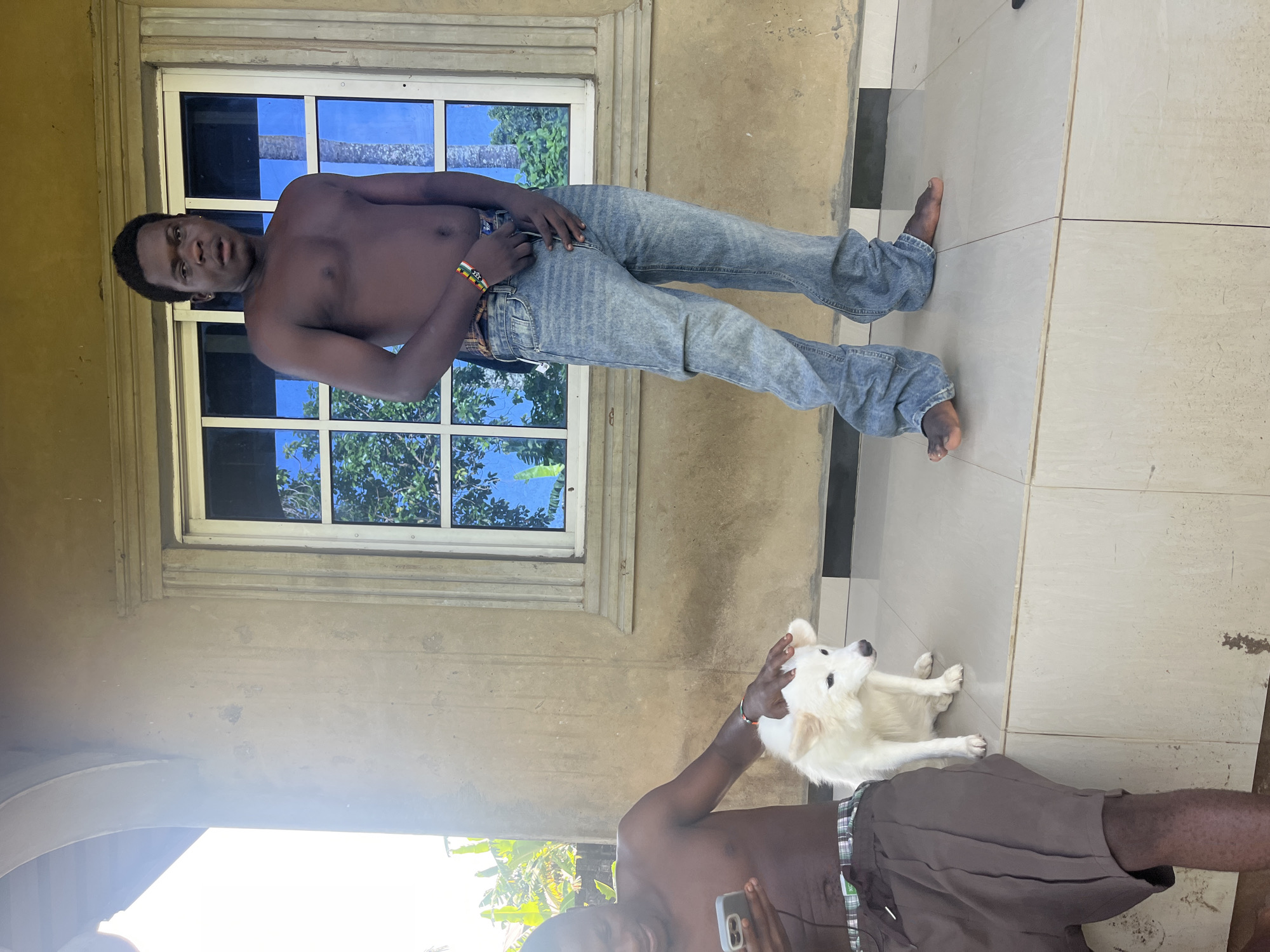 A medium shot shows two shirtless men and a white dog on a tiled floor in Ikot Ekpene, Nigeria.
The man nearest the camera is partially in the shot, with the camera capturing his torso and legs. He wears brown shorts and is holding a smartphone in his left hand. He has his right hand on the dog’s head.
The second man stands closer to the window, wearing light-blue jeans.
The floor is comprised of large, light-colored tiles with dark inlays. A portion of a light-colored exterior wall is visible, featuring a window. The window is multi-paned. Outside, there are green trees and a blue sky.