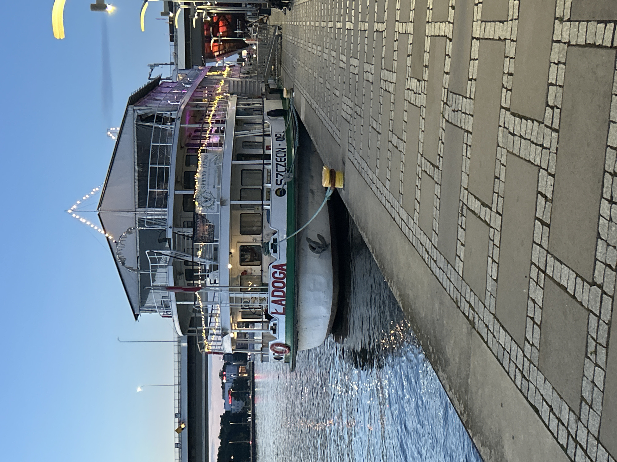 This is a wide-angle, eye-level, outdoor shot taken in Szczecin, Poland, during twilight. The scene features a large, white paddlewheel boat docked alongside a stone-paved promenade. The boat, named "ŁADOGA," is illuminated with strings of festive lights, some of which are pink and others yellow. The boat has multiple decks and appears to be a venue for events, with a closed-off upper deck covered by a tent-like structure. The name "SZCZECIN - OPL" is also visible on the side of the boat. The water of the Odra River is calm and reflects the lights. In the background, a bridge spans the river, and the lights of the city are visible on the far bank. The promenade is paved with rectangular stone tiles and smaller cobblestones arranged in a patterned fashion. A yellow bollard is present on the dock. Two people are partially visible sitting on a bench on the right side of the promenade, near some lights.