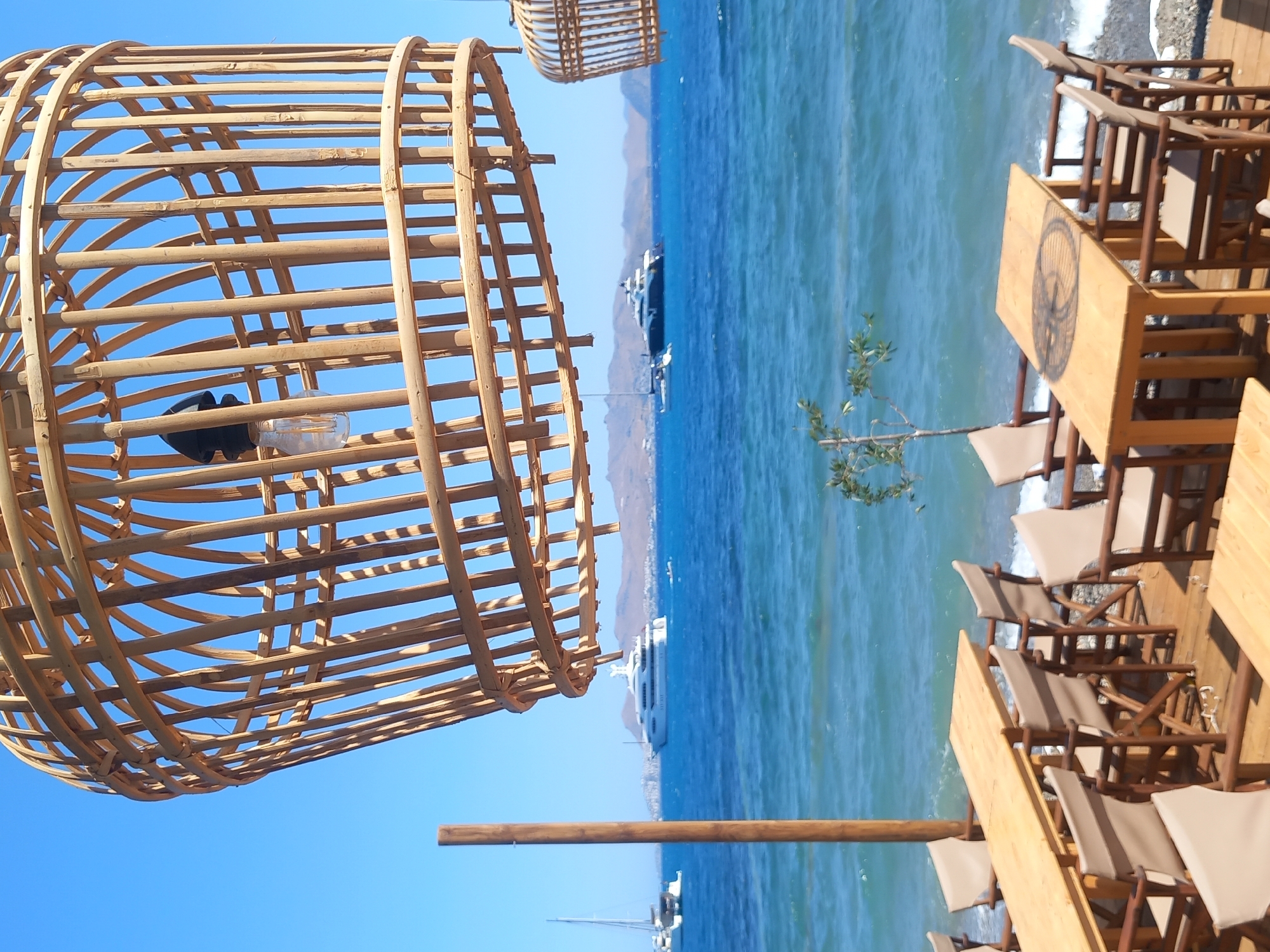 This is a photograph taken outdoors, likely at a beachfront restaurant or cafe. In the foreground, a large, intricate wicker light fixture hangs from above, obscuring the upper portion of the image. Below the light fixture, several wooden tables and chairs are arranged on a wooden deck. The tables are simple, light-colored wood, and the chairs appear to be made of wood with fabric seats and backs.
Beyond the seating area, the sea stretches out, a vibrant blue under a clear sky. Waves are visible, gently lapping at the shore. In the distance, several yachts and sailboats are anchored or cruising on the water. The coastline is lined with hills or mountains. The overall impression is one of a serene and picturesque setting, likely in a warm, sunny climate.
While the exact location within the city of Kos, Greece, cannot be definitively determined without further context, the presence of yachts and the coastal landscape are consistent with the island's geography and popular tourist destinations.