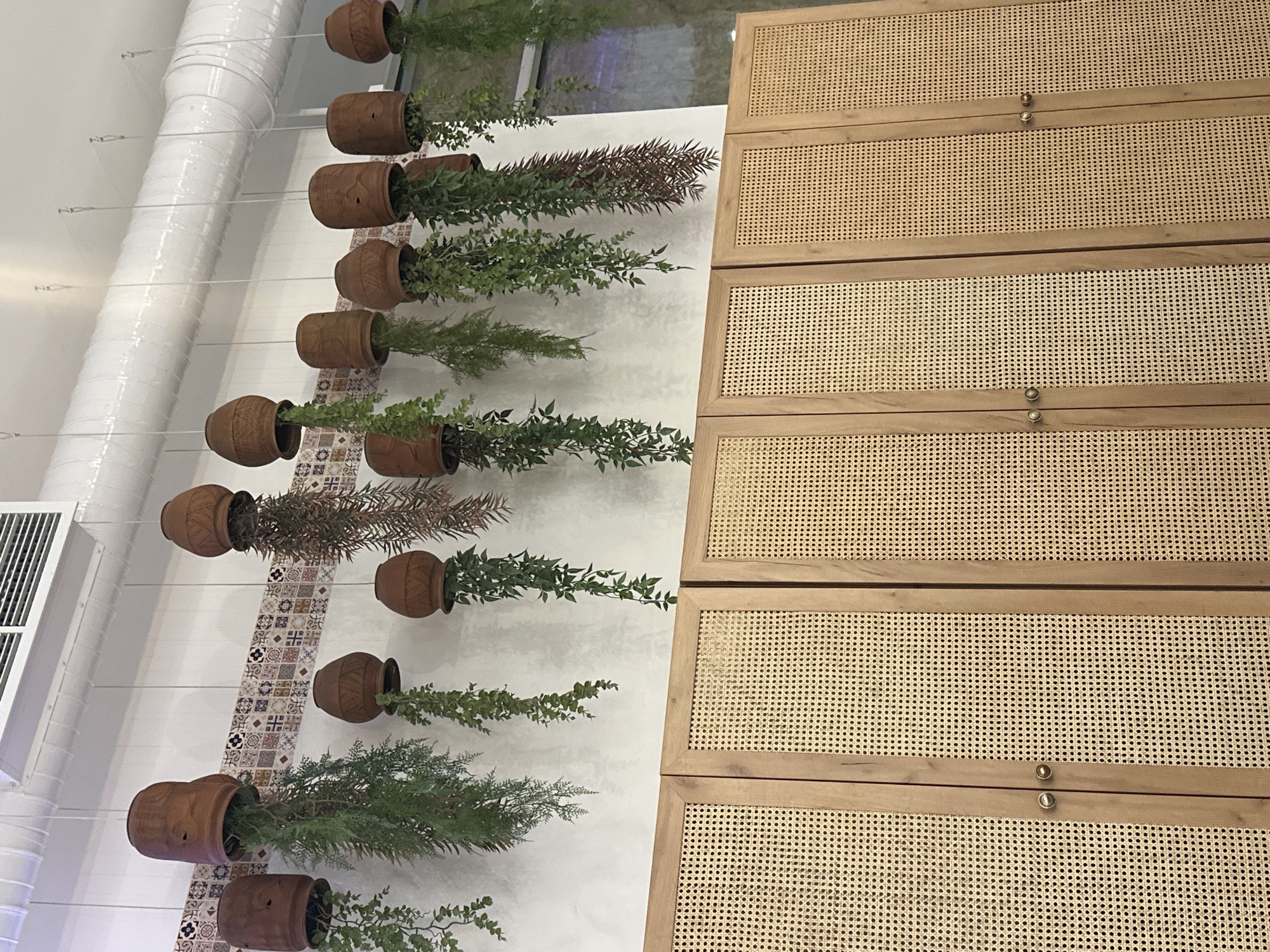 The image depicts an interior scene with a focus on decorative elements. On the left, terracotta pots are suspended from the ceiling, each containing trailing green plants. These pots are arranged in a vertical line against a white wall adorned with a patterned tile strip. To the right, a cabinet or dresser features a light wood frame with woven cane panels. The overall aesthetic suggests a modern, bohemian-inspired design. The location in Saint Petersburg, Russia is not identifiable from the image provided.
