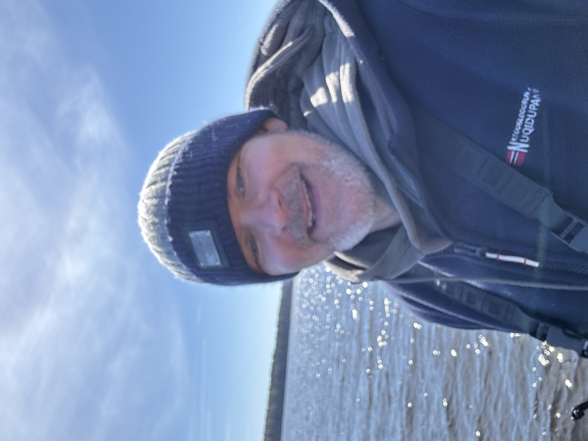 A man with a light beard and wearing a dark blue and gray beanie and a dark hooded jacket is posing for a selfie. The jacket has a logo that appears to read "Nuqidupan." The background consists of a body of water reflecting sunlight and a strip of trees on the horizon under a clear blue sky with faint clouds. The location is not ascertainable as Aleksandrovka, Russia, with the provided information.