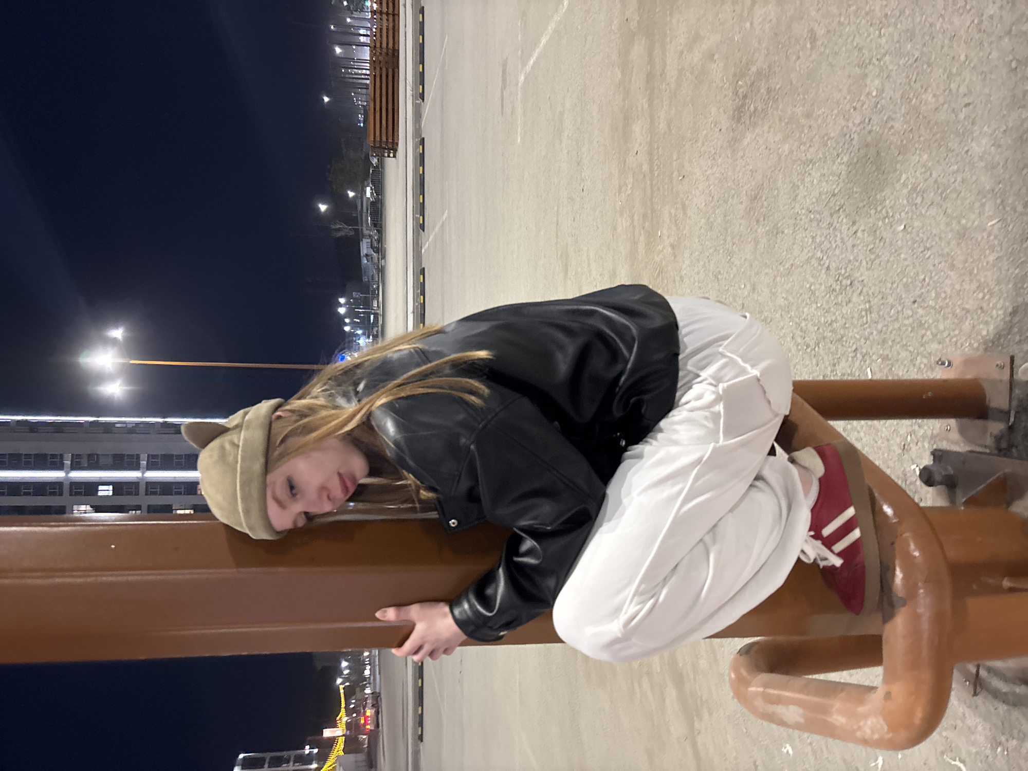 The image depicts a young woman in an urban setting, likely in Perm, Russia. She is leaning against a metal structure, possibly part of a public space installation or fitness equipment. She wears a beige hat with cat ears, a black leather jacket, and white pants. One of her feet, clad in a red and white Adidas sneaker, is resting on the structure. The background consists of a parking lot or large paved area, buildings, and streetlights. The time of day appears to be dusk or night.