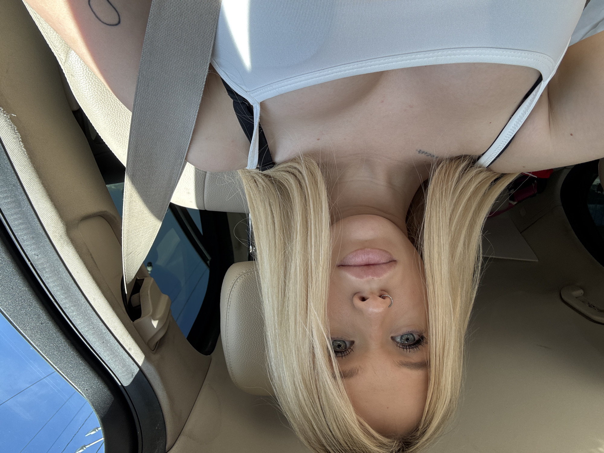 This is a selfie taken from the perspective of someone sitting in the passenger seat of a car. The subject is a young woman with long blonde hair, blue eyes, and a nose ring. She is wearing a white tank top. The interior of the car is visible, including a beige leather seat and a seatbelt. The background shows a bright blue sky and some power lines, suggesting an outdoor setting on a sunny day. There is no information in the image to suggest a location in Saint Petersburg, Russia.
