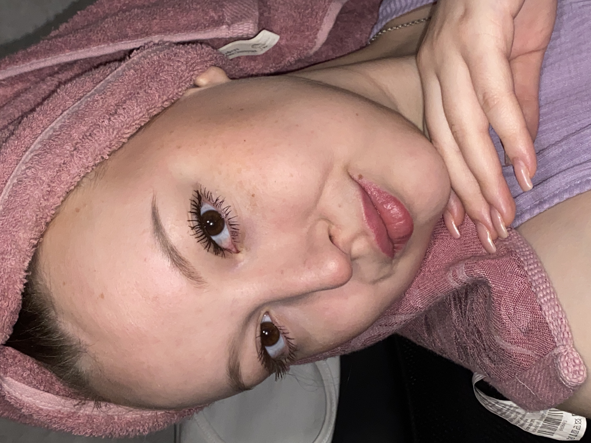 The image displays a young woman lying down and looking towards the viewer, with her head tilted, causing the frame to appear rotated counter-clockwise. Her light brown hair is encased in a textured, dusty pink or mauve towel, suggesting she has recently washed it. She possesses fair skin, with visible freckles, and striking dark brown eyes enhanced by mascara. Her eyebrows are well-defined, and her full lips are slightly parted, adorned with a glossy, pinkish-red product. She wears a relaxed, subtly playful expression, possibly a slight smirk or pout.
Her right hand is visible near her neck and shoulder, showcasing neatly manicured nails painted in a natural, light nude shade. She is dressed in a light lavender or purple ribbed top, complemented by a delicate silver chain necklace. The background is dark and out of focus, indicating an indoor setting.
Visible text includes a white fabric tag on the pink towel, clearly showing "H&M" and care symbols. Another white fabric tag, partially visible near the bottom right, displays a barcode and the number "690893", along with additional care instructions. The scene captures a moment of casual relaxation, likely after a shower or bath. This image was taken in Chaikovsky, Russia.