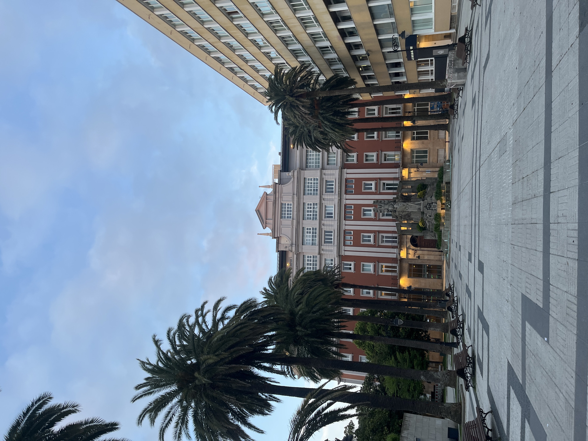 The image shows a street scene in La Coruña, Spain. Tall buildings flank a pedestrian square paved with gray and white stones. A row of palm trees lines one side of the square, interspersed with benches and lampposts. The buildings exhibit a mix of architectural styles, with one appearing modern and another with a red-brick facade and white window frames. The sky is overcast, suggesting either dawn or dusk. No people are visible in the immediate scene.