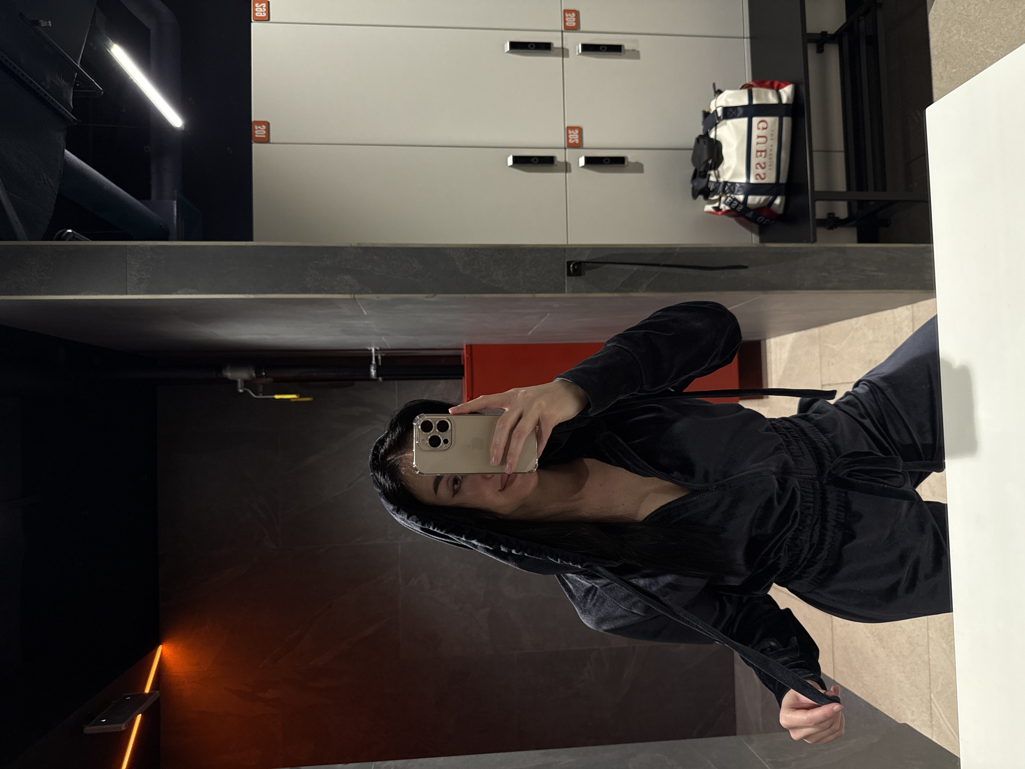The image shows a woman in a dark velvet sweatsuit taking a selfie in what appears to be a locker room or bathroom. The background includes lockers with numbers visible, a Guess brand duffel bag, and what looks like a shower area or wall with orange lighting. The location is likely a fitness facility or gym somewhere in the city of Krasnoyarsk, Russia.