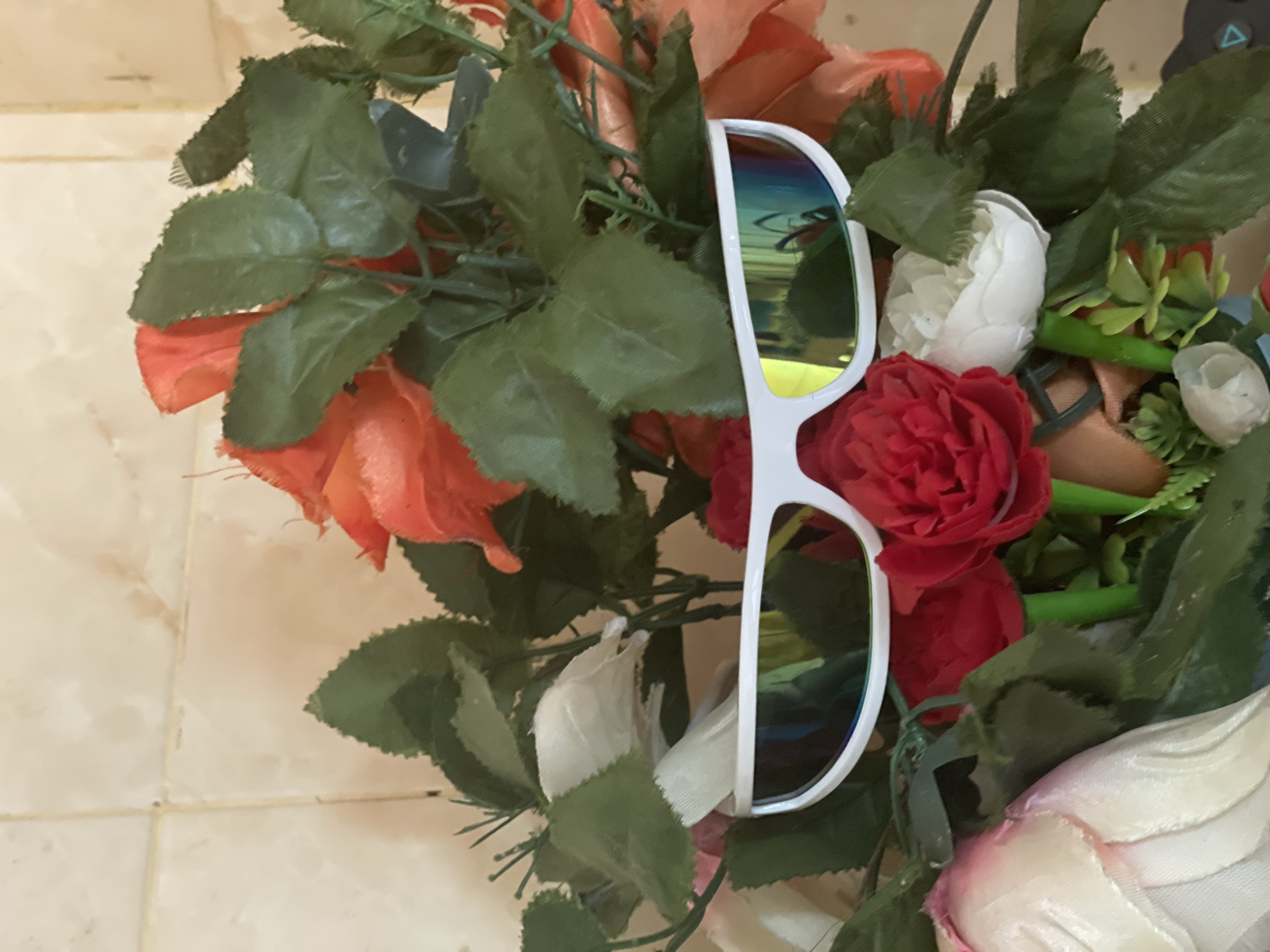 In Ado, Nigeria, a close-up, eye-level shot captures a bouquet of artificial flowers and a pair of white futuristic sunglasses placed in the foreground, obscuring the floral arrangement. The sunglasses, with their reflective, iridescent lenses, create a striking contrast against the vibrant red, orange, and white faux roses and green leaves. The background features a tiled wall, suggesting an indoor setting, possibly a home or commercial space. The lighting is even, indicating a well-lit environment.