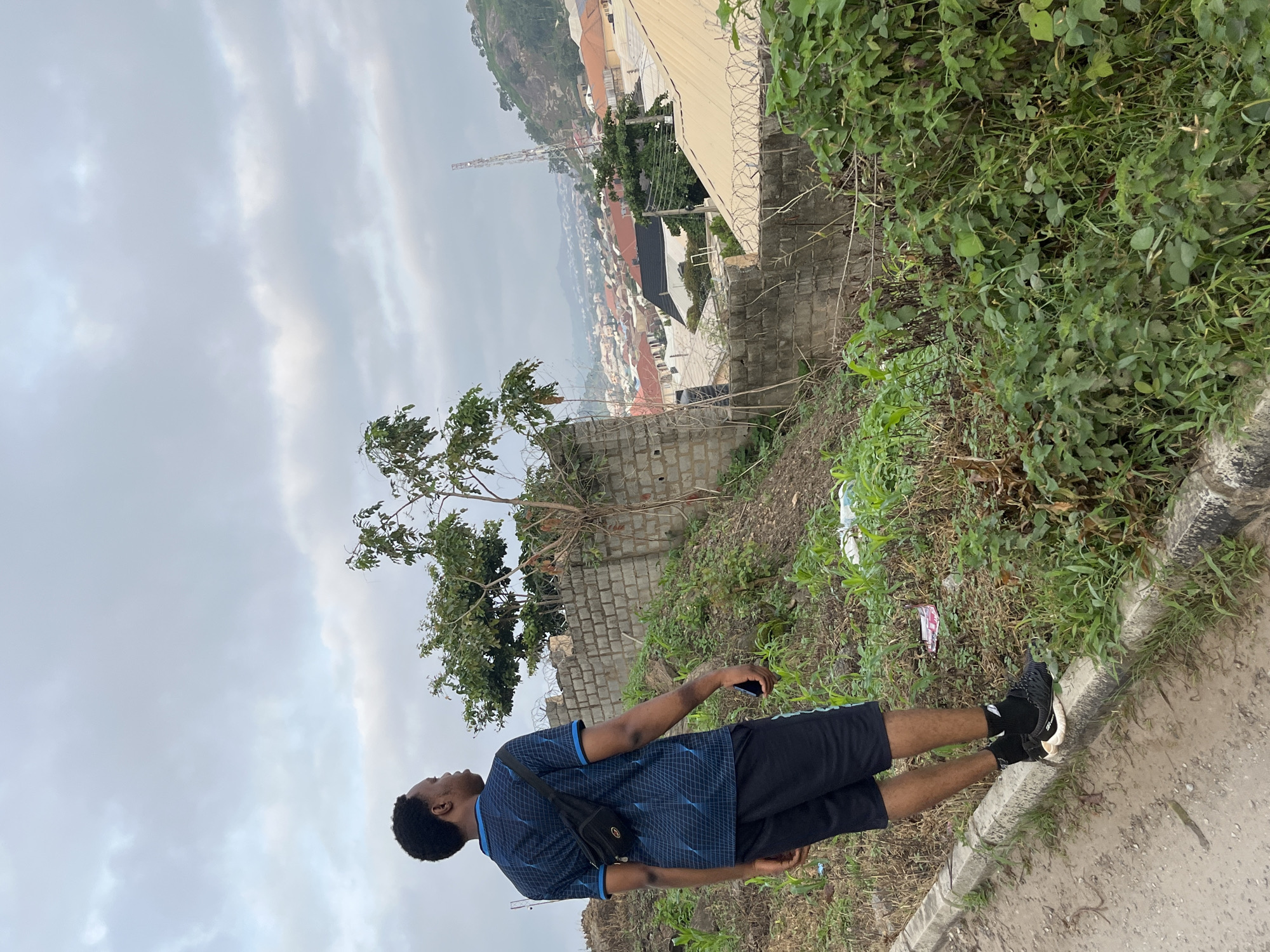 A young Black man is standing on a dirt road, looking out over a city. He is wearing a blue t-shirt with a geometric pattern, black shorts with blue trim, black socks, and black athletic shoes. He has a black fanny pack slung across his chest. The man is standing on a curb next to a steep, overgrown embankment. In the background, a city spreads out across a hilly landscape. Buildings with various colored roofs are visible, and a tall transmission tower stands on a distant hill. The sky is overcast. This scene is consistent with a view from an elevated area in Abuja, Nigeria, possibly in a residential district overlooking the main city.