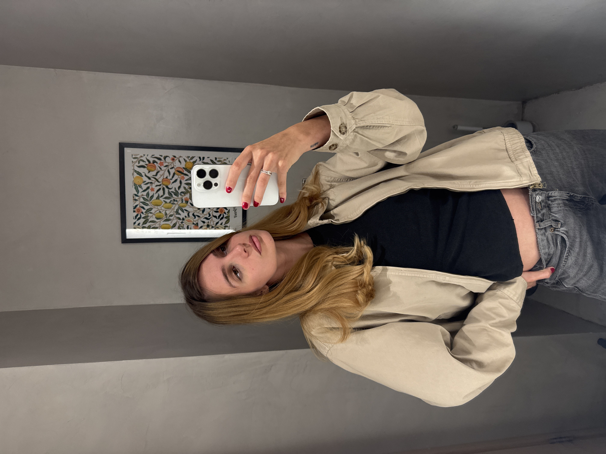 A woman is taking a selfie in what appears to be a bathroom. The walls are a uniform, light grey color. A framed print with a pattern of leaves and citrus fruit hangs on the wall behind her. The woman has long, blonde hair with subtle waves. She is wearing a black top, a beige jacket, and grey jeans. Her fingernails are painted red, and she has a ring on her finger. The phone she is using to take the picture is white. A roll of toilet paper is visible in the background. There are no indicators in the image suggesting the location is Yerevan, Armenia.
