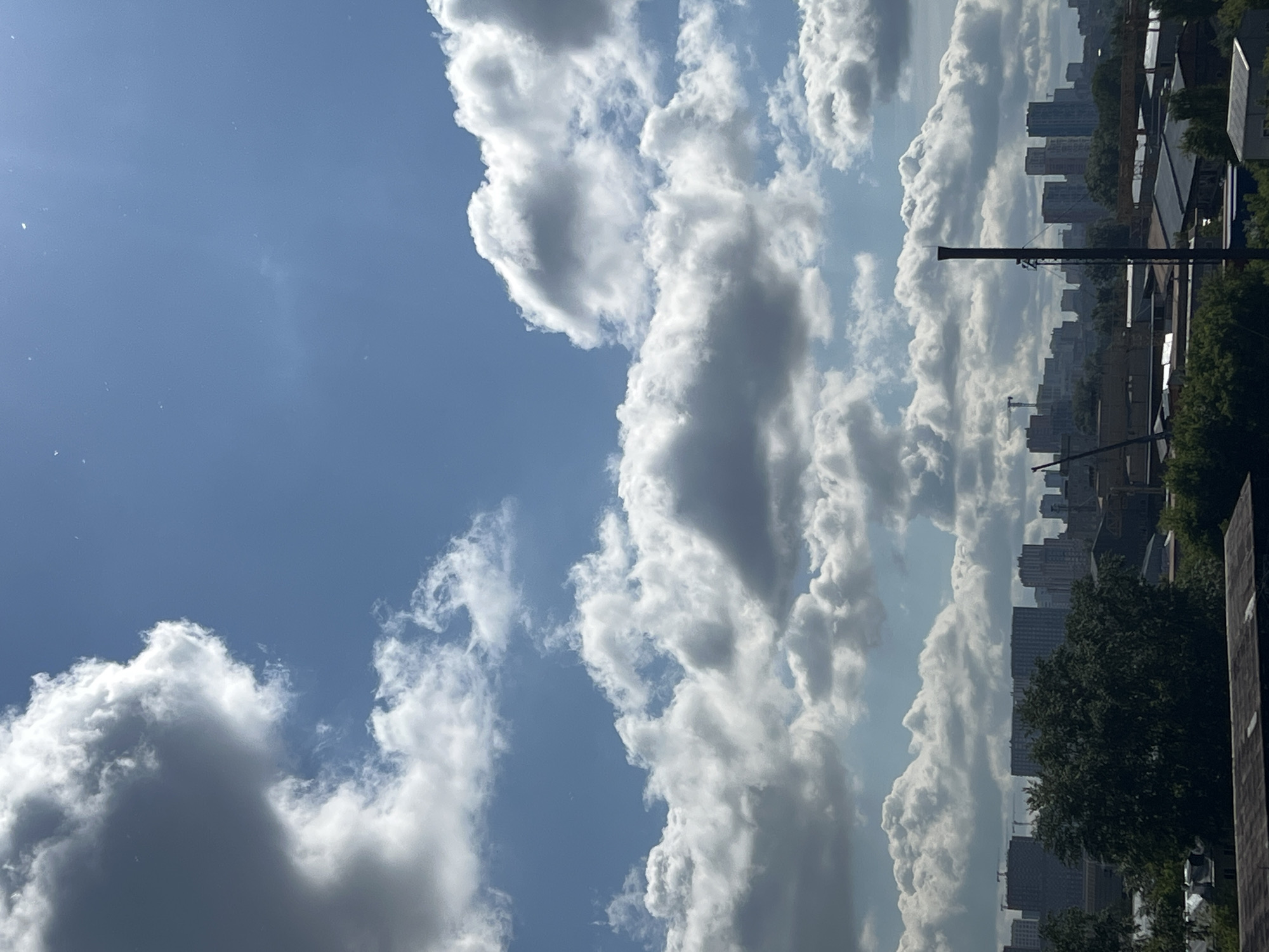 The image captures a view of a city skyline under a sky filled with dynamic clouds. In the foreground, a dark roofline is visible. Above this, a dense green tree dominates the mid-ground, partially obscuring a collection of buildings that form the city's silhouette. These buildings vary in height and architectural style, with some appearing to be under construction, indicated by cranes. A prominent dark, slender industrial chimney or structure stands tall amidst the buildings in the mid-right portion of the frame. The sky is a vibrant blue, punctuated by large, white, and grey cumulus clouds, some casting shadows on the buildings below. The sun is visible in the upper right, creating a lens flare effect and illuminating the scene. The overall impression is that of an urban landscape on a partly cloudy day, possibly during daylight hours. Based on the architectural styles and general urban development, this scene is consistent with an industrial or residential area within Yekaterinburg, Russia.