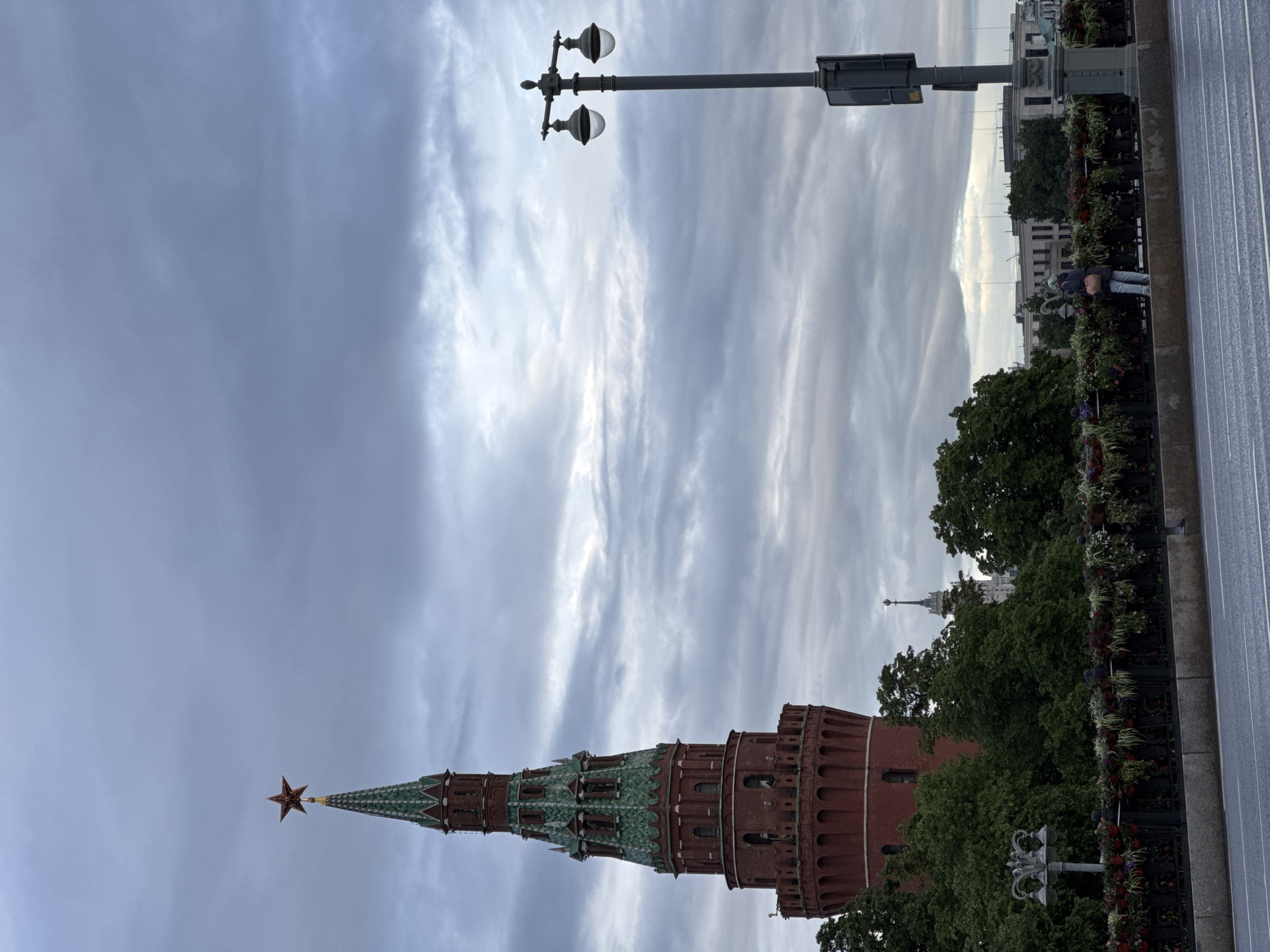 The image shows a view of the Kremlin in Moscow, Russia. The tower with a red star on top is the dominant feature, rising against a cloudy sky. In the background, other buildings are visible behind trees, including what appears to be a skyscraper. The foreground shows a street with a decorative fence and flowers. A lamppost stands near the street. A person is visible in the background, standing on the sidewalk.